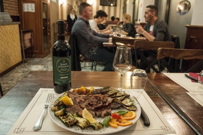 Piatto di carne e verdure alla griglia con bottiglia di vino sul tavolo di un ristorante. Persone che cenano sullo sfondo.