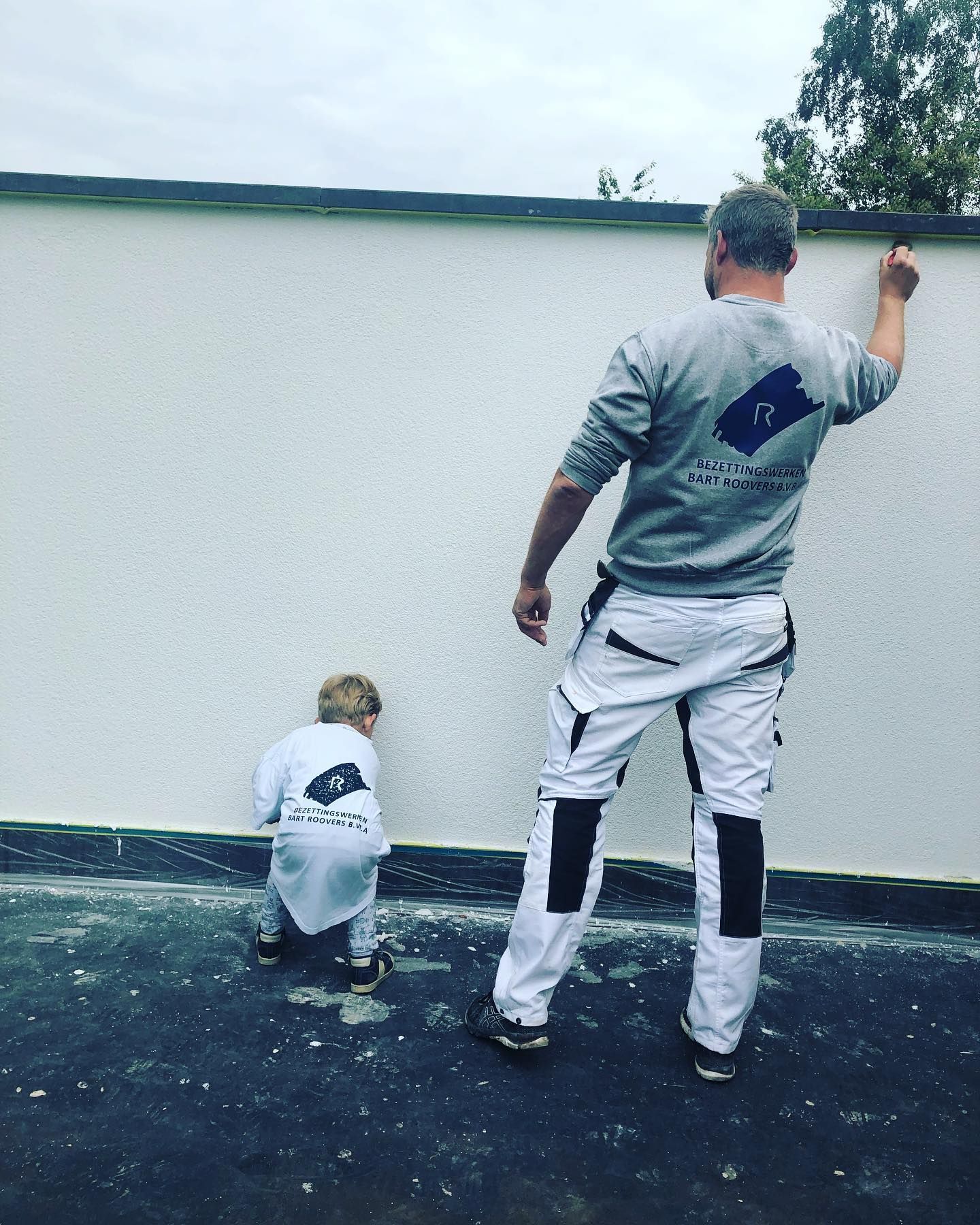 Een volwassene en een kind in bijpassende sportkleding staan buiten bij een witte muur.