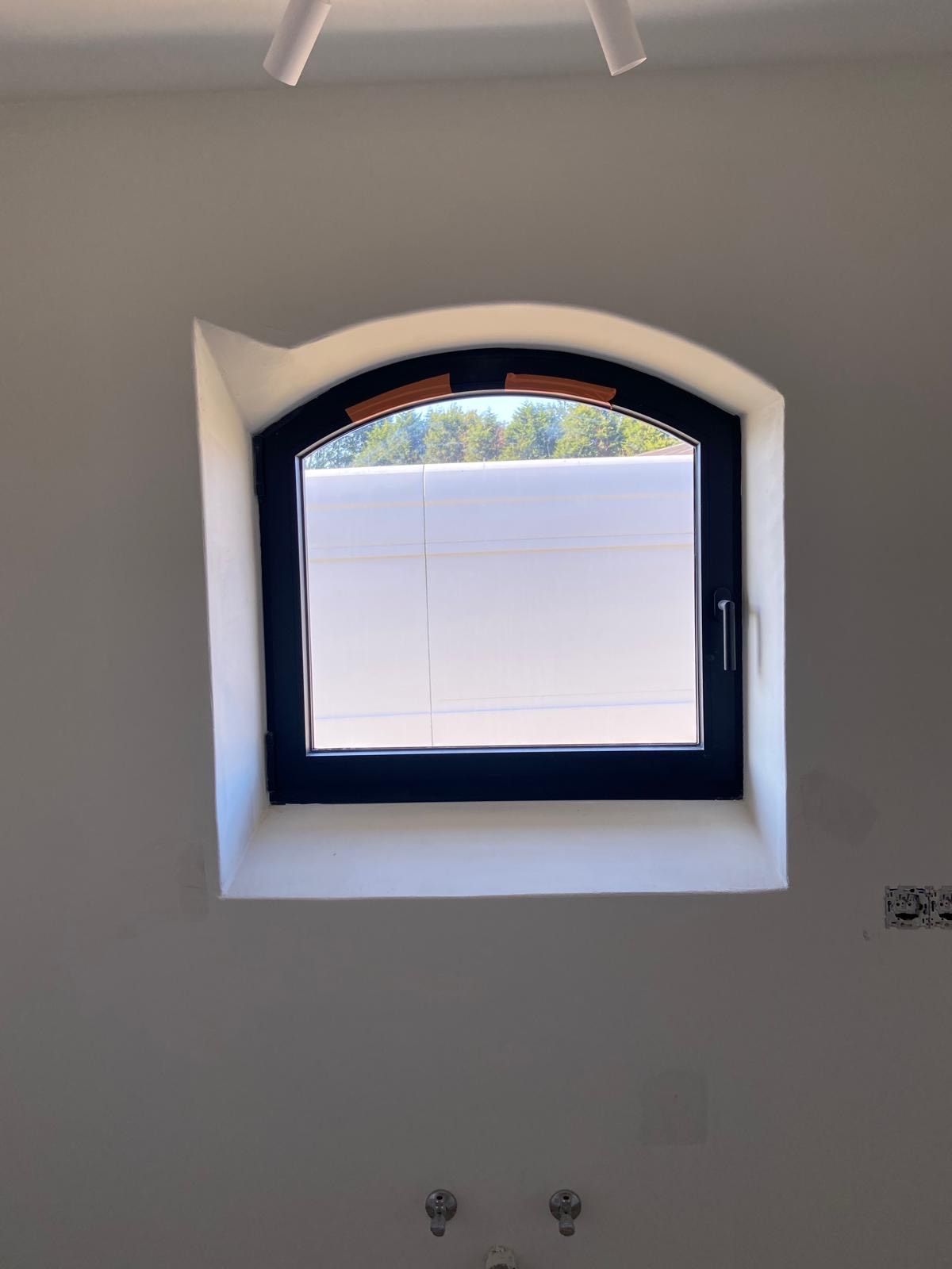 Klein boogvormig raam met een donker frame in een witte muur, gedeeltelijk aan de buitenkant bedekt door een wit paneel.