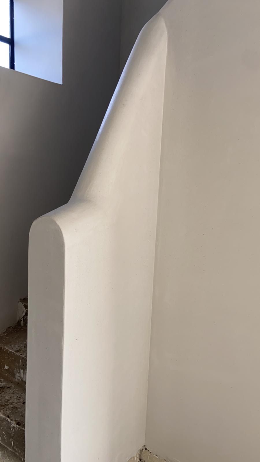 Witte, gebogen trapmuur naast een donker trappenhuis en een klein raam.