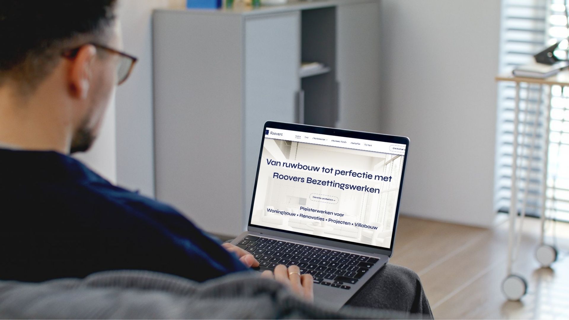 Een persoon zit op een bank in een lichte woonkamer en gebruikt een laptop, met een presentatiedia op het scherm.