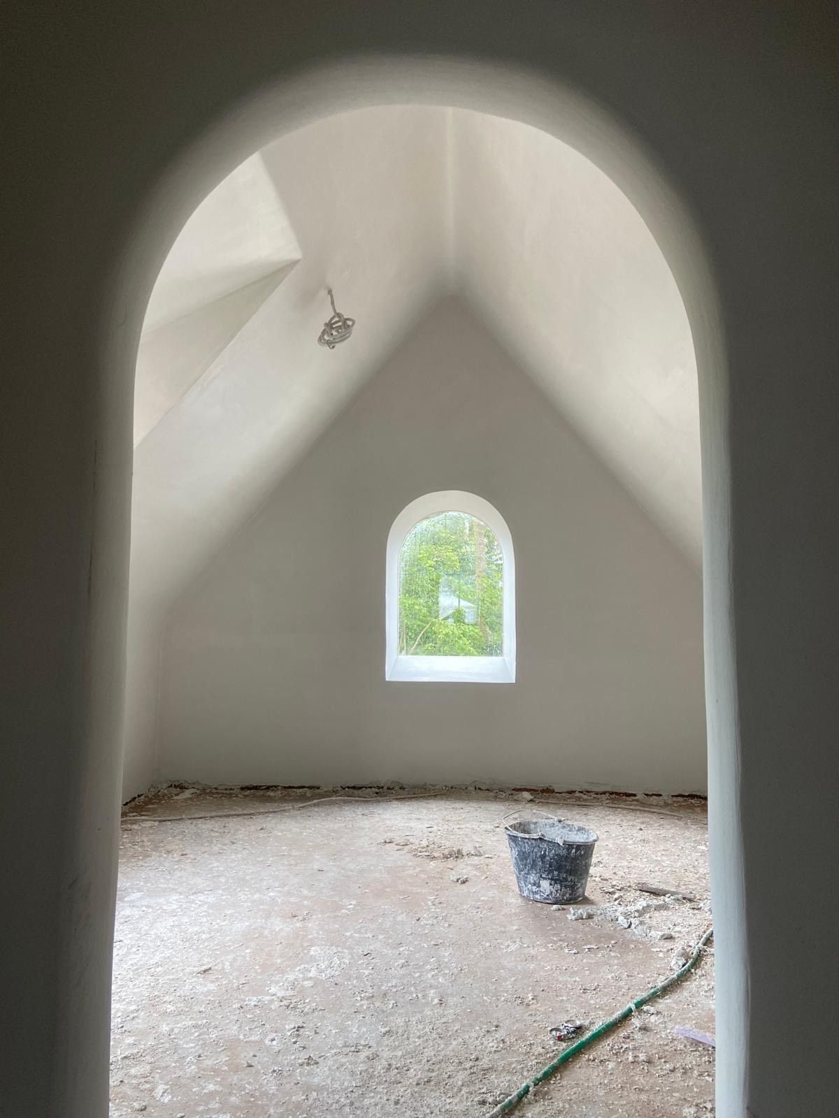 Lege, witte, gewelfde ruimte in aanbouw met een klein boogvormig raam en een emmer op de vloer.