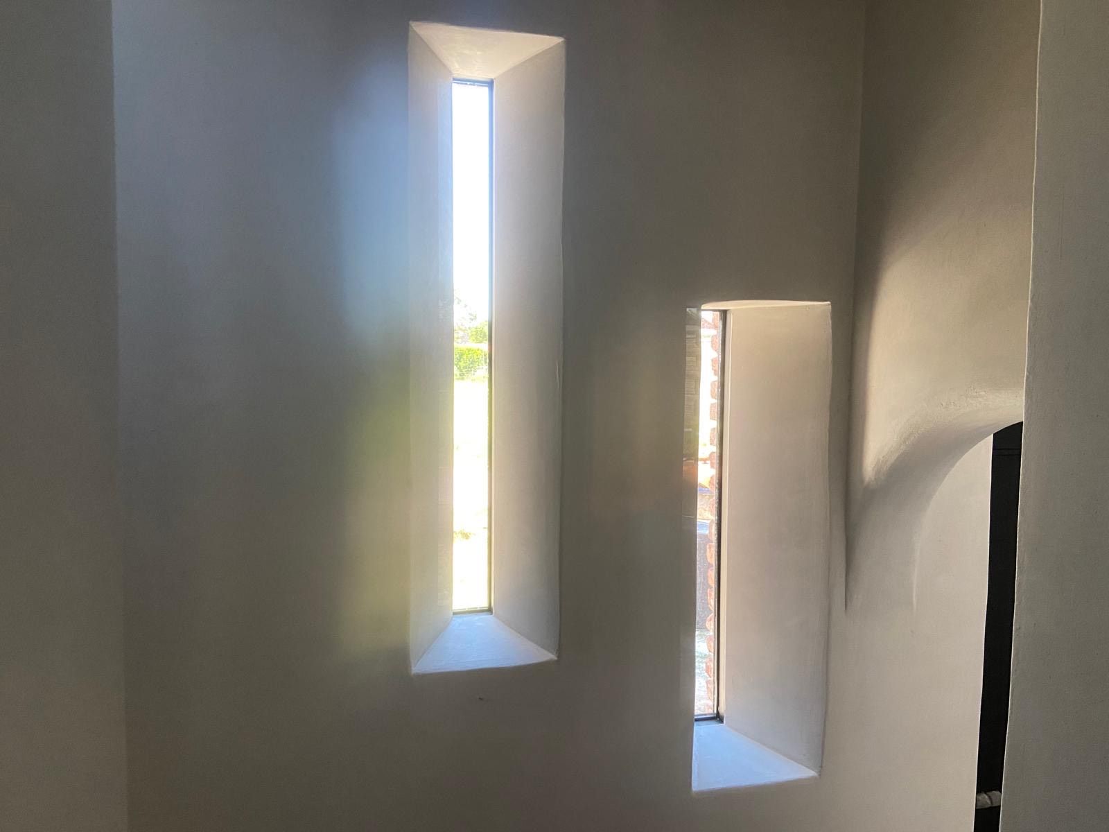 Twee smalle, verticale ramen in een grijze binnenmuur, met zonlicht en een glimp van groen buiten.