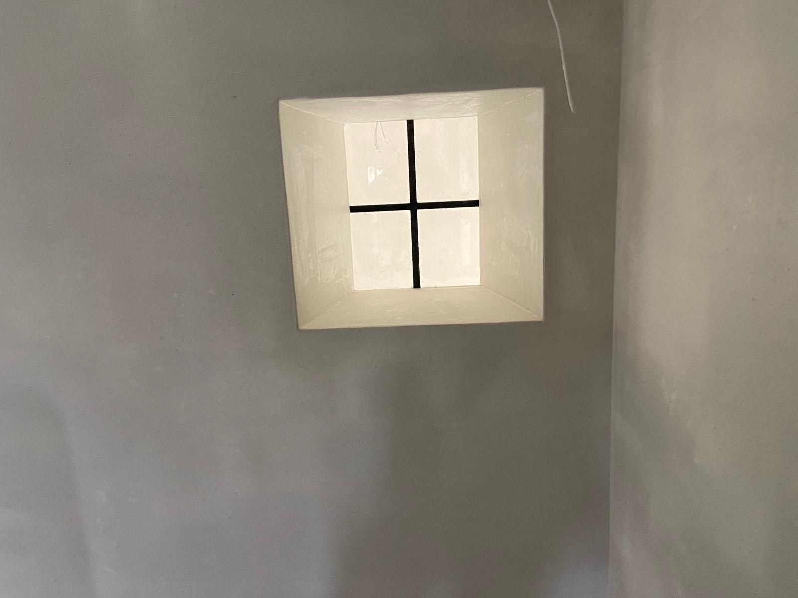 Klein vierkant raam met een zwart kruisvormig kozijn in een grijze muur.