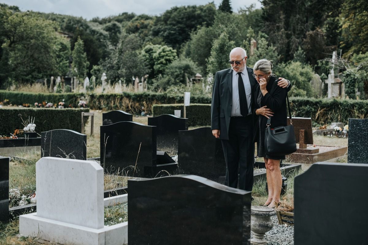 Man in suit comforts woman in black dress at a cemetery, graves in view, trees in background.