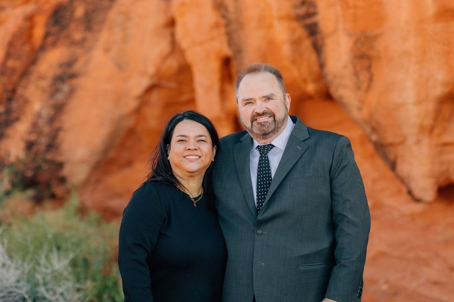 Woman in olive dress and man in navy suit, standing together. Both are smiling outdoors.