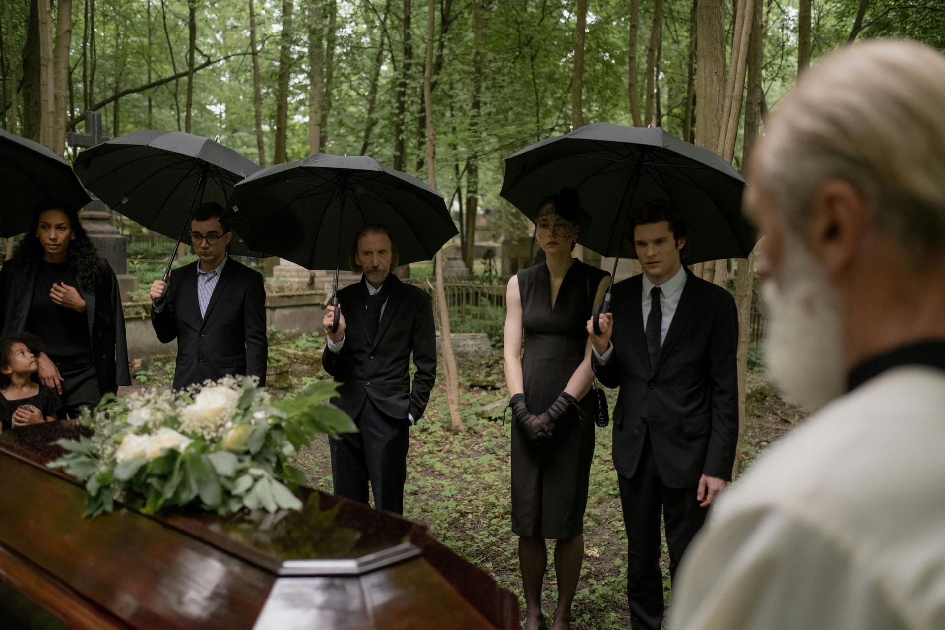 People in black attire stand at a casket in a cemetery, holding umbrellas.