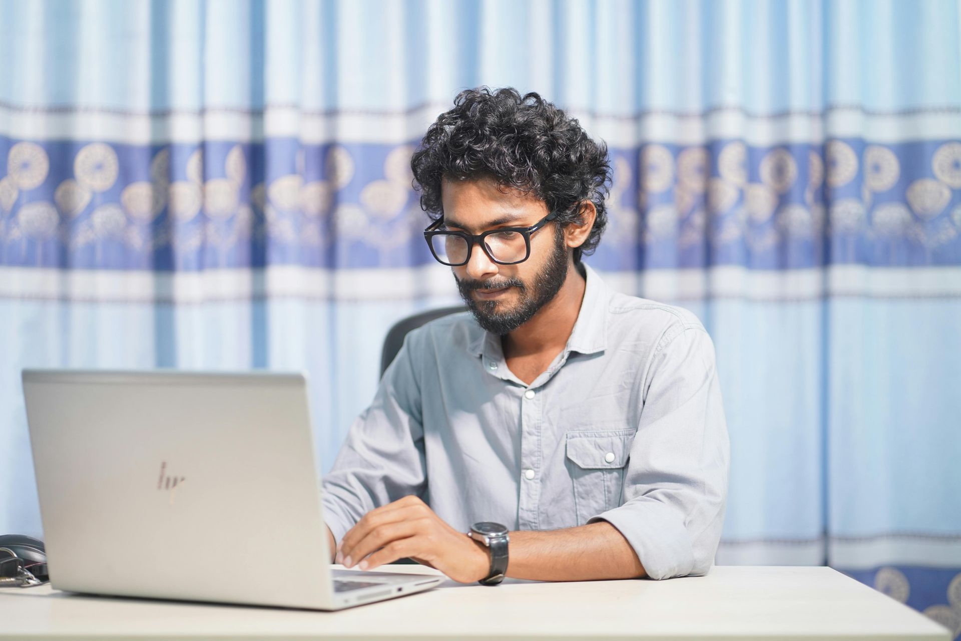 Man with glasses working on a laptop at a desk, light blue shirt, blue curtain background.