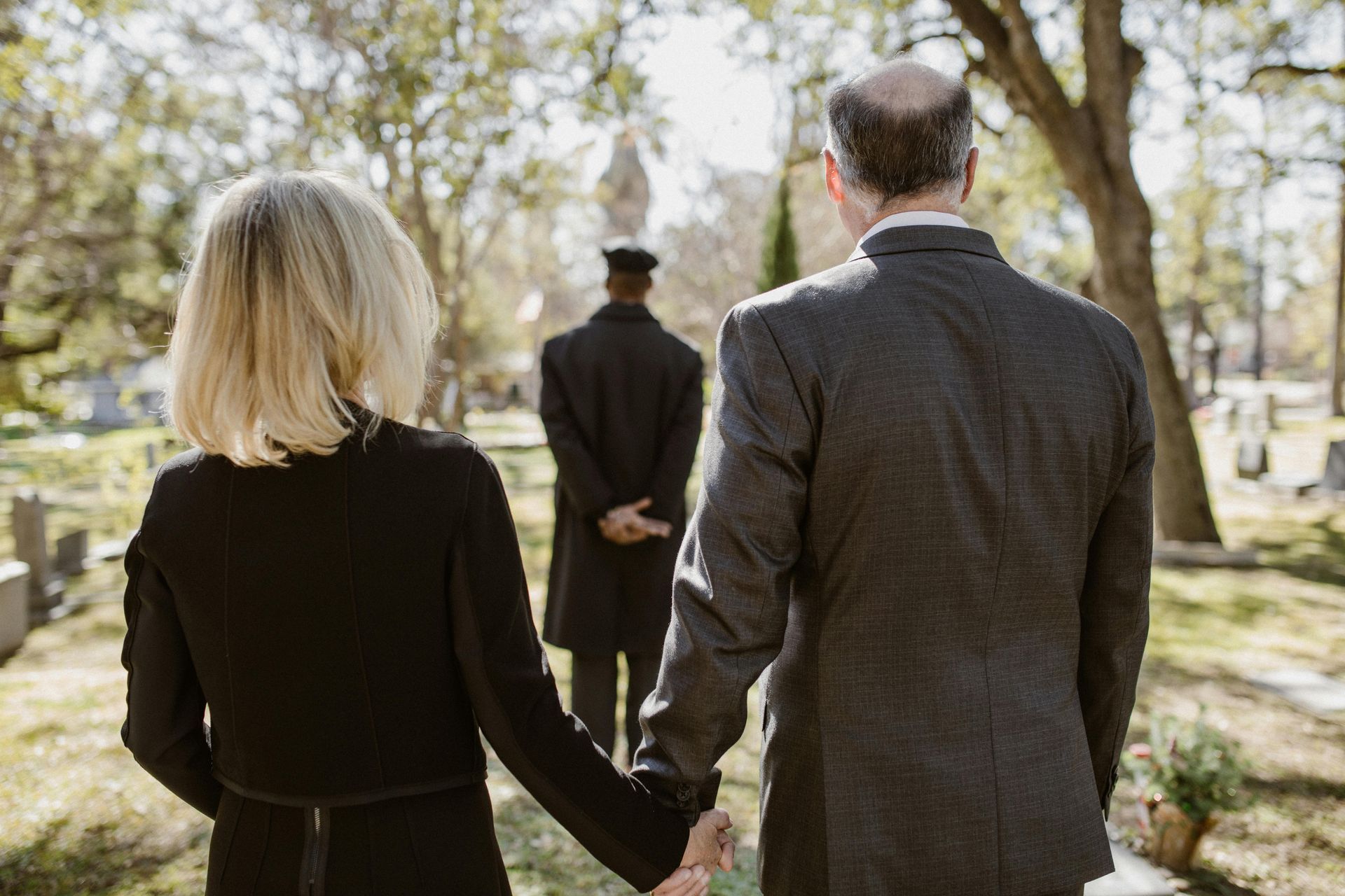 A couple holding hands walks towards a person in black in a cemetery, trees and tombstones visible.