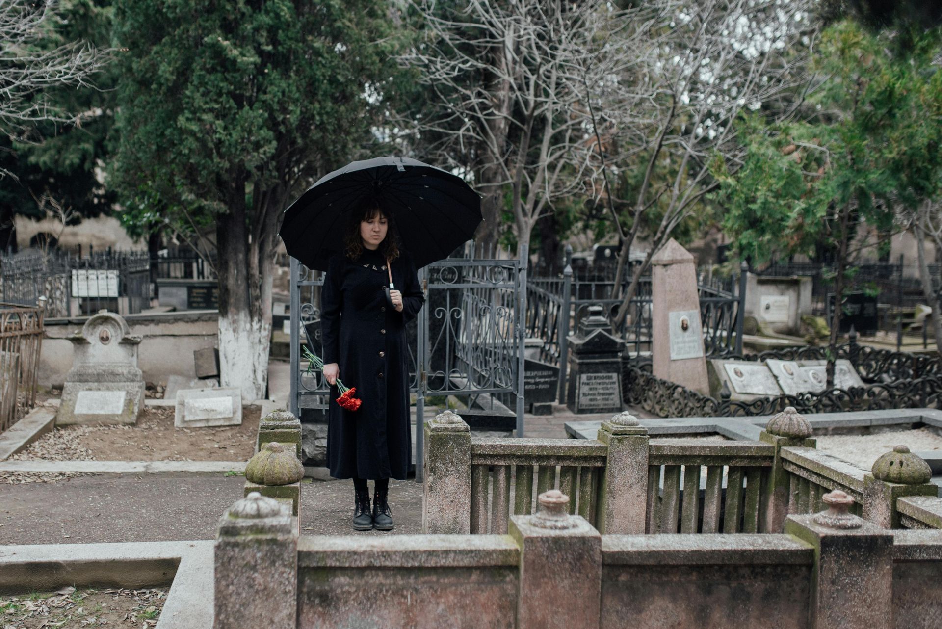Woman in black coat holds an umbrella and flower, standing in a cemetery with tombstones.
