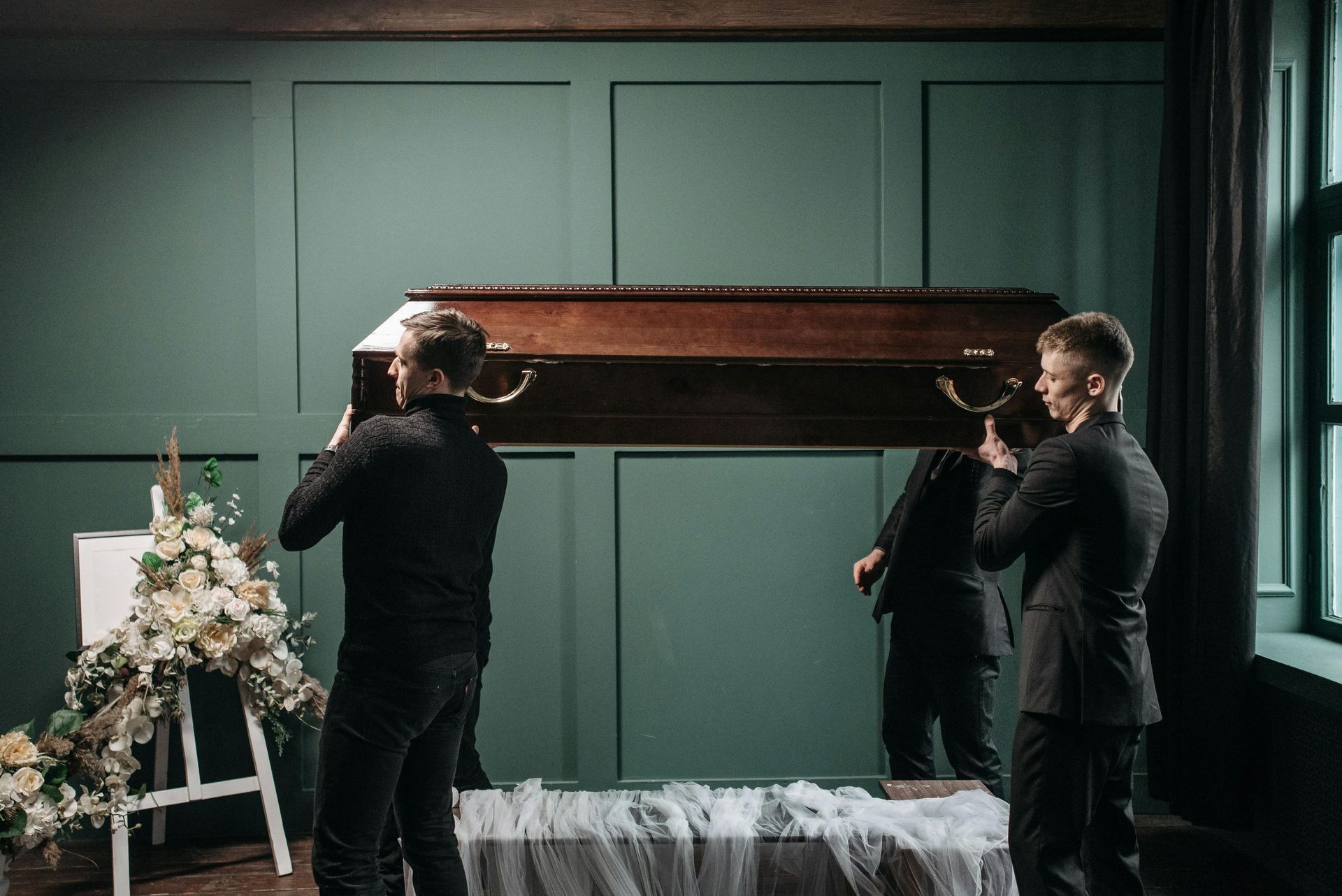 Men carrying a casket at a funeral. Dark suits, green wall, floral arrangement, and a window.