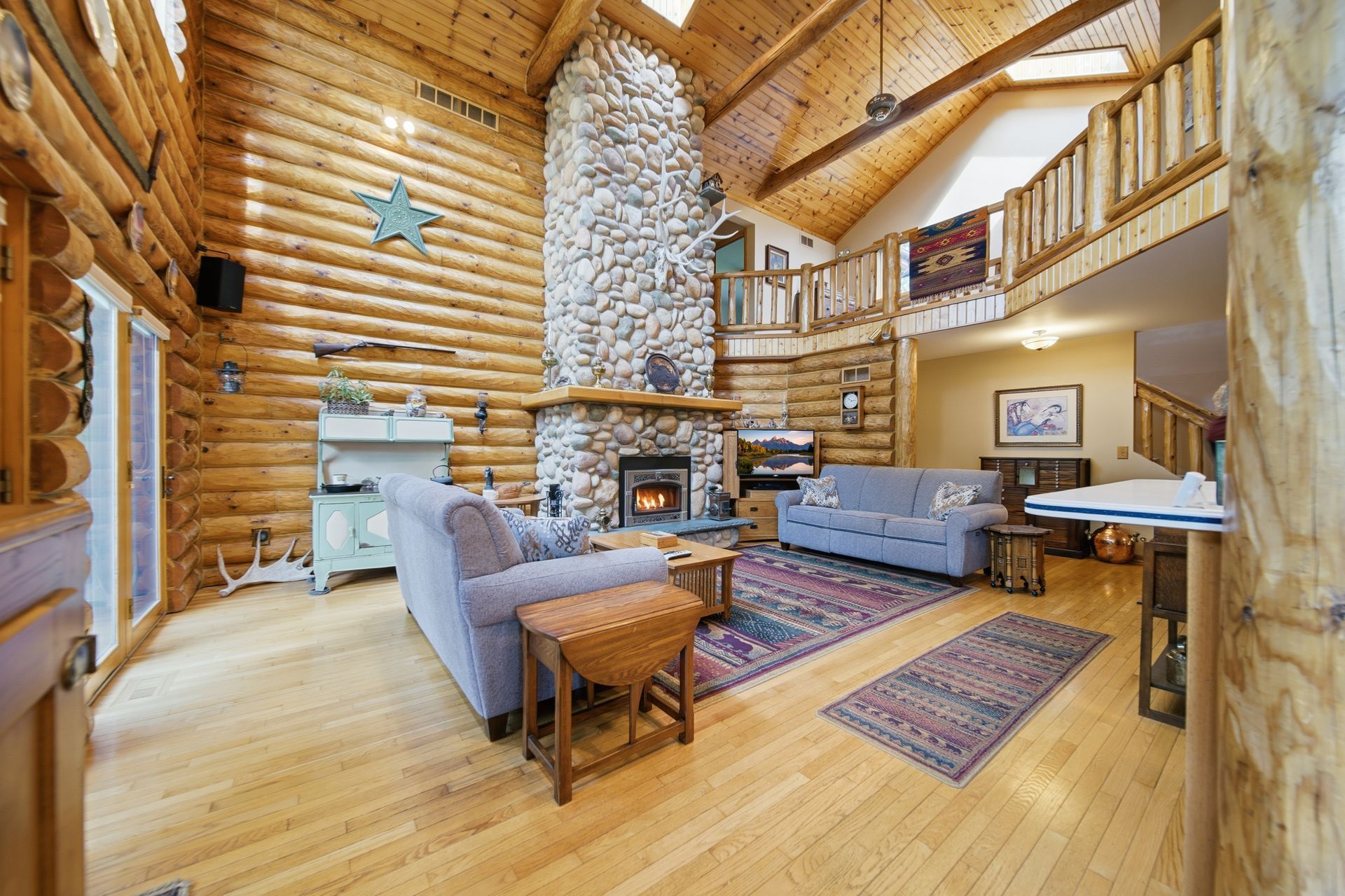 Double-height log home living room with a stone chimney and loft railing