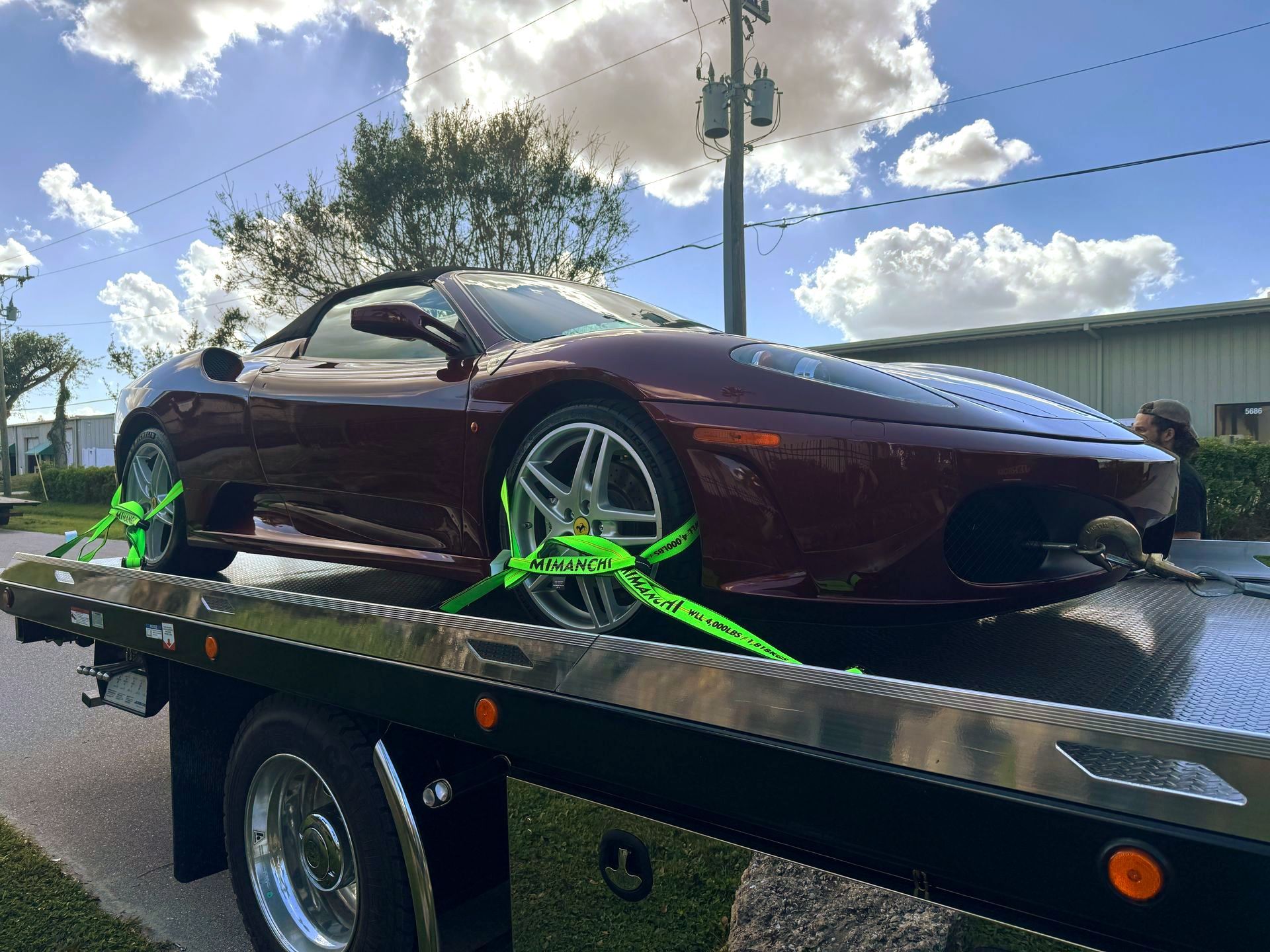 A dark red Ferrari convertible is securely strapped to a flatbed tow truck outdoors on a sunny day.