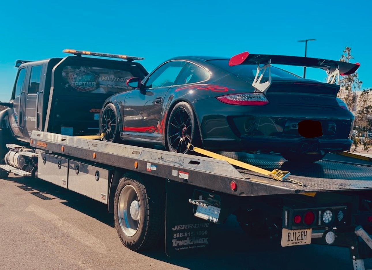 A black Porsche sports car with a large rear wing is secured on the flatbed of a tow truck.