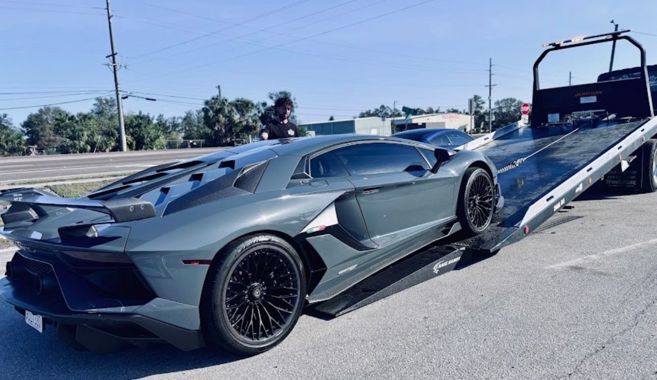 A gray Lamborghini Aventador being loaded onto a flatbed tow truck in an outdoor parking lot on a sunny day.