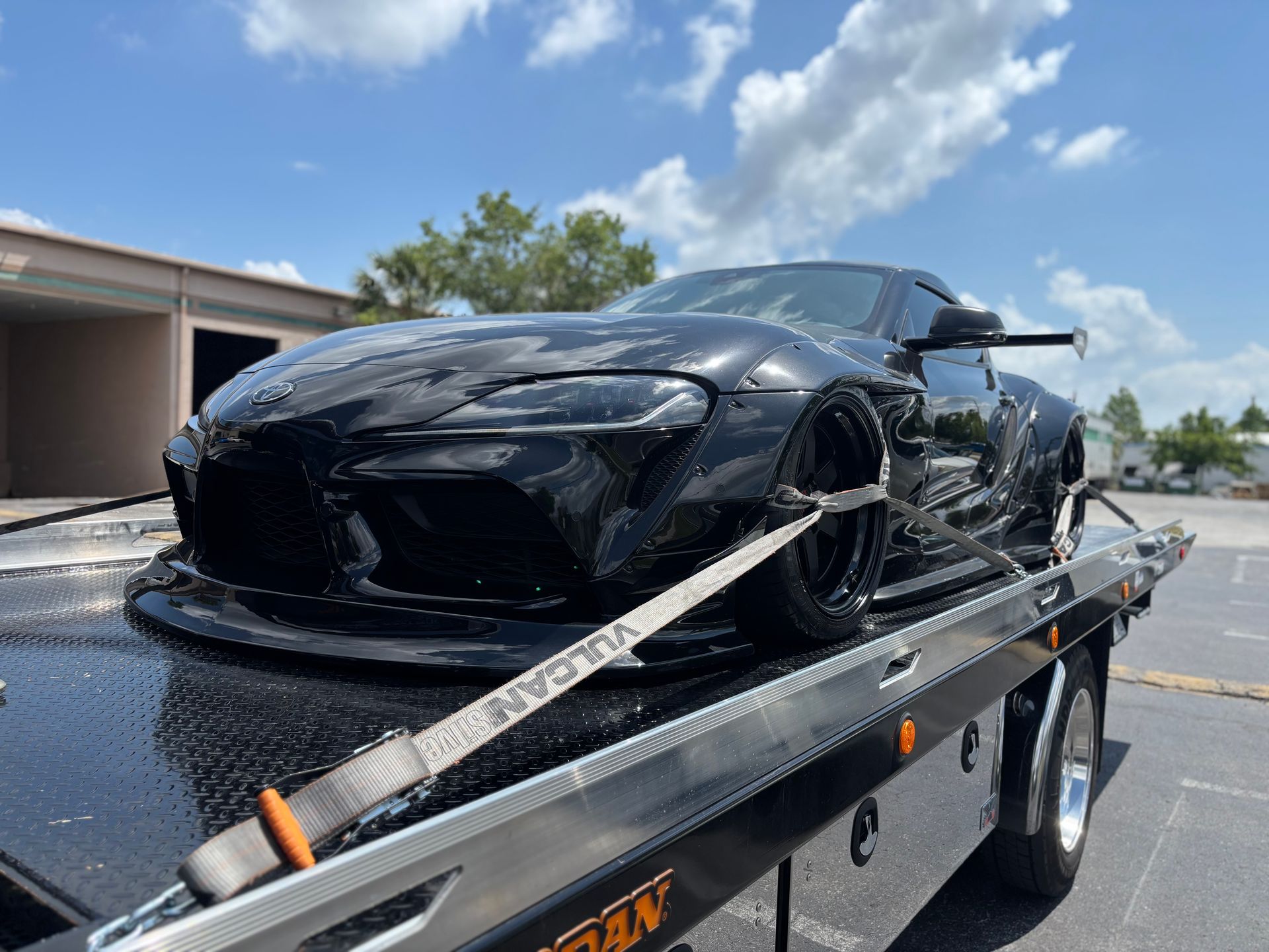 A black Toyota Supra strapped down and secured on a flatbed tow truck on a sunny day.