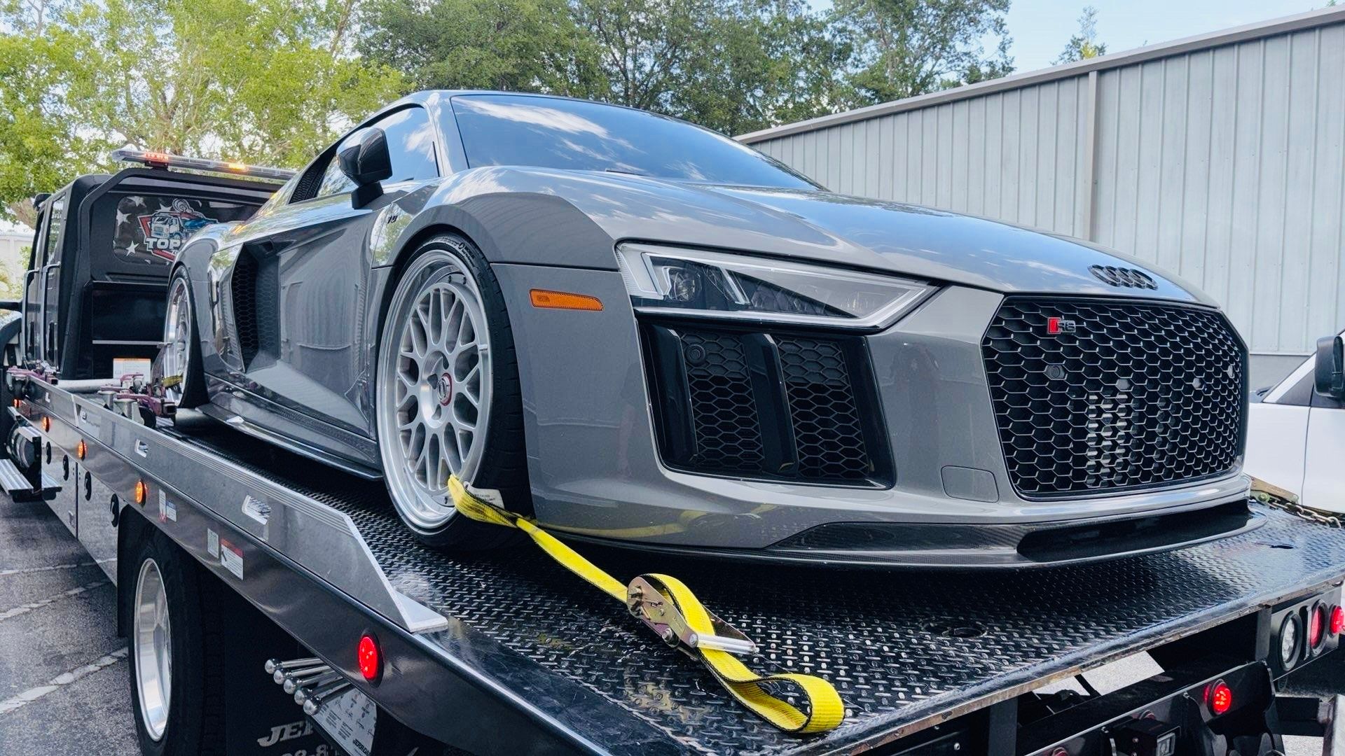 A gray Audi R8 sports car secured with yellow tie-down straps on a flatbed tow truck.