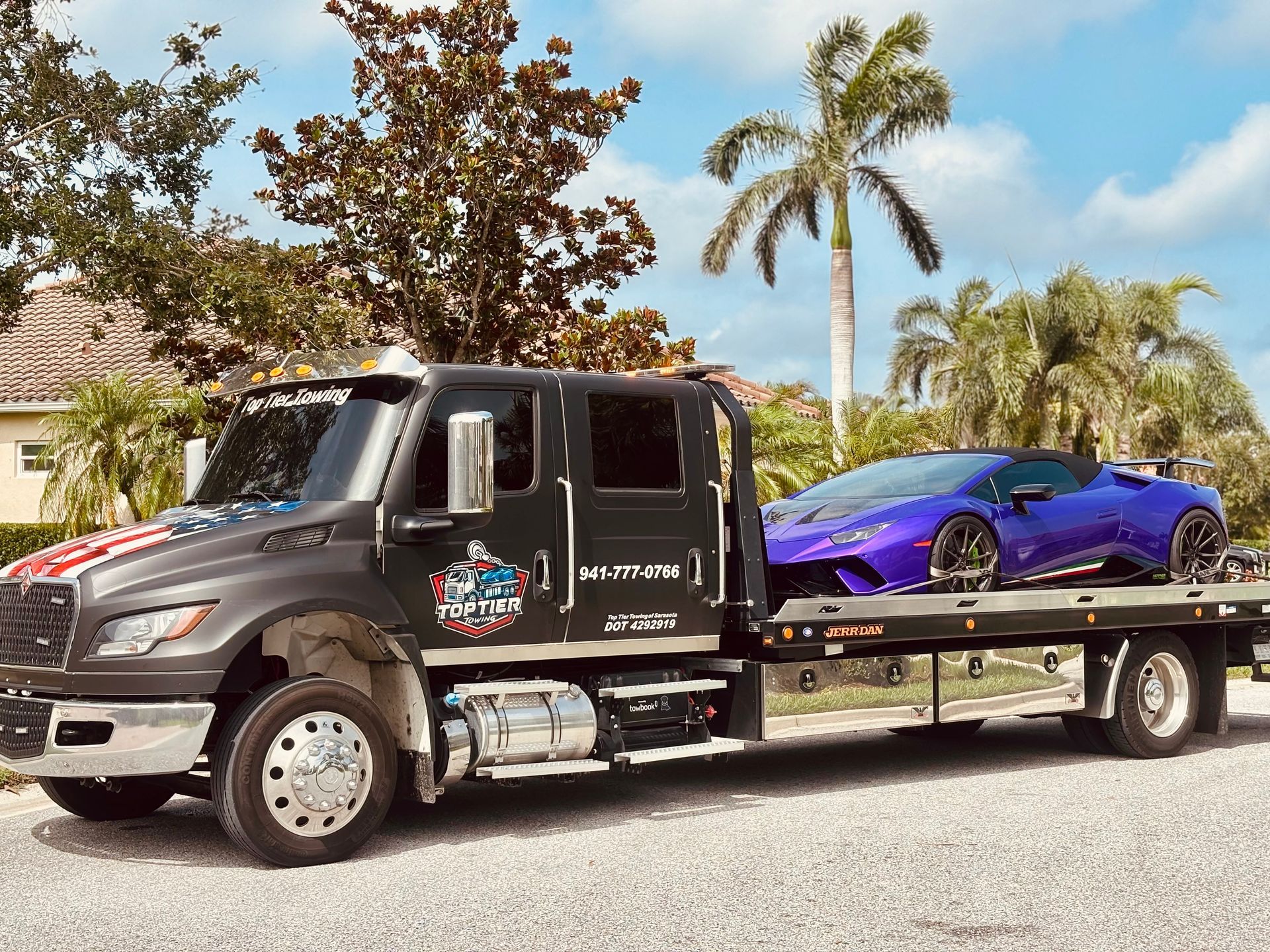 A black flatbed tow truck carries a purple Lamborghini sports car against a backdrop of palm trees and a residential home.