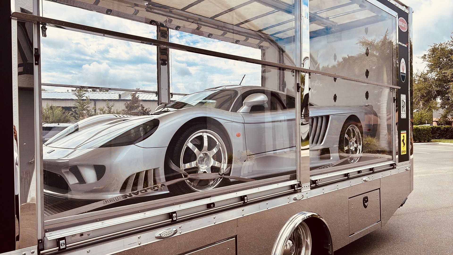 A white Audi R8 with a colorful, geometric racing wrap and large rear spoiler strapped to the back of a flatbed tow truck.