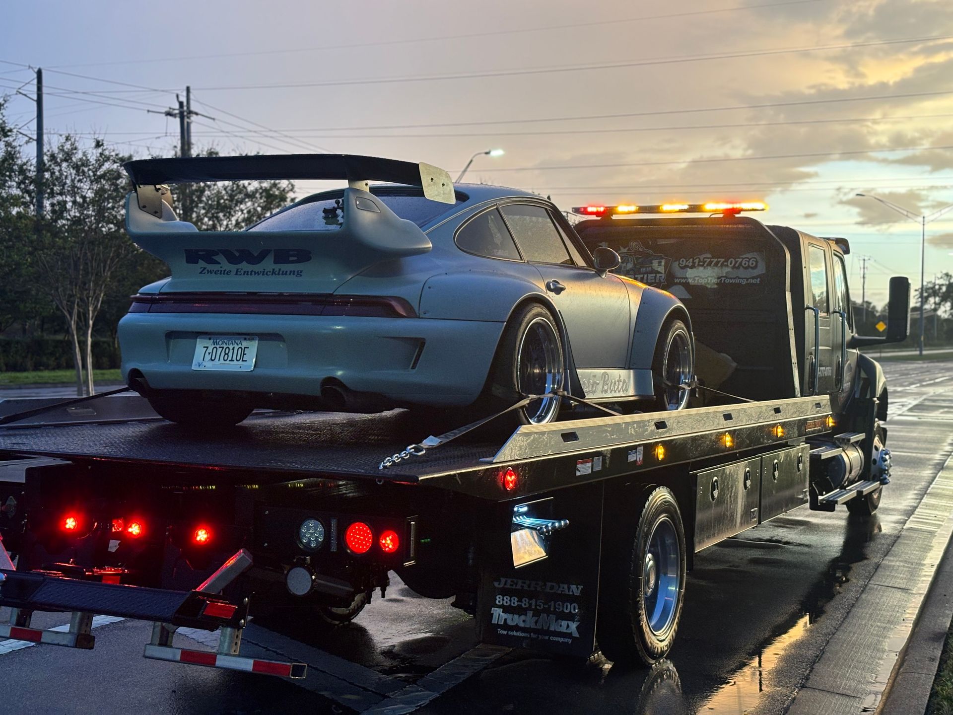A matte grey RWB Porsche 911 with a large rear wing is loaded onto the flatbed of a tow truck at sunset.