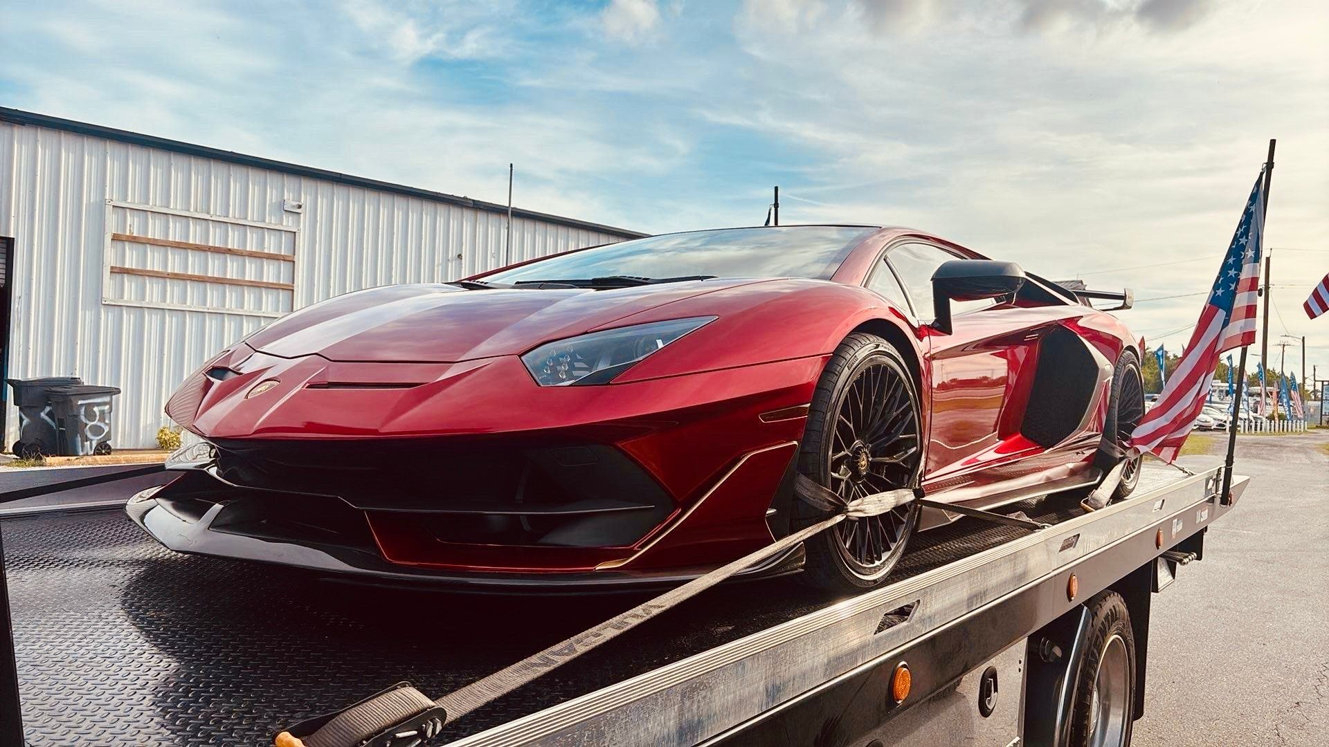 A metallic red Lamborghini on a flatbed tow truck with an American flag attached to the back.