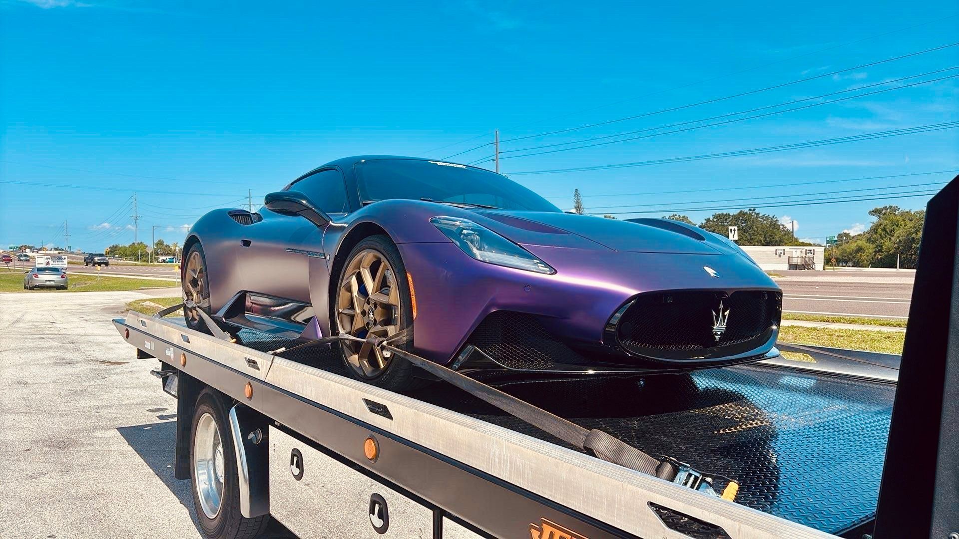 A purple Maserati MC20 sports car secured on the flatbed of a tow truck parked in an outdoor lot.