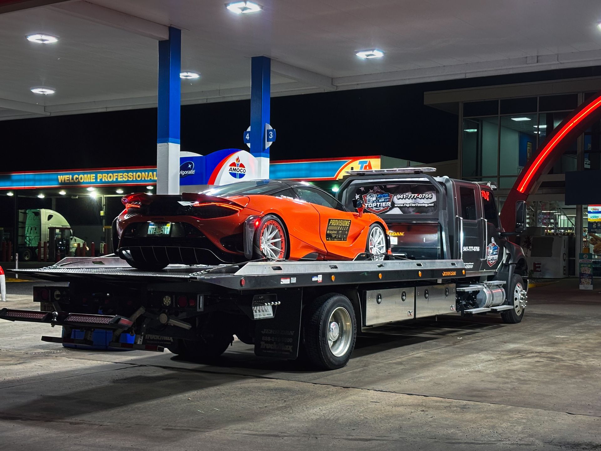 A bright orange McLaren supercar is loaded onto the flatbed of a tow truck at a gas station at night.