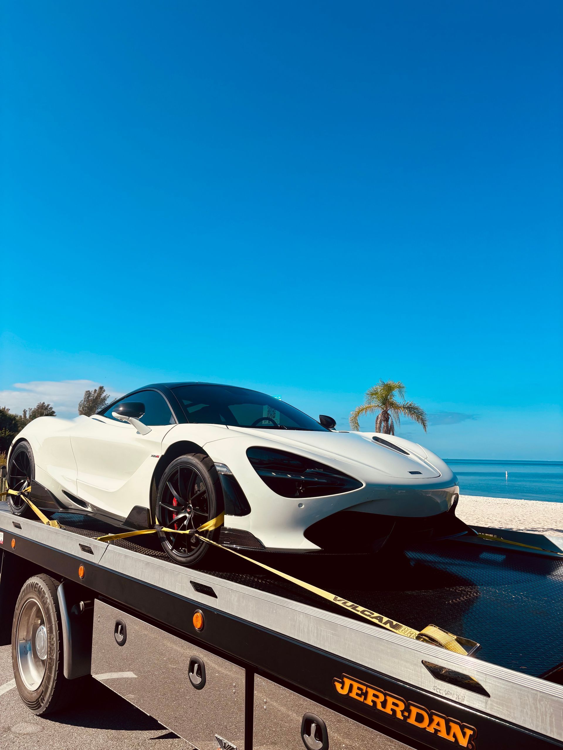 A white McLaren sports car strapped onto a Jerr-Dan flatbed tow truck parked near a beach under a clear blue sky.