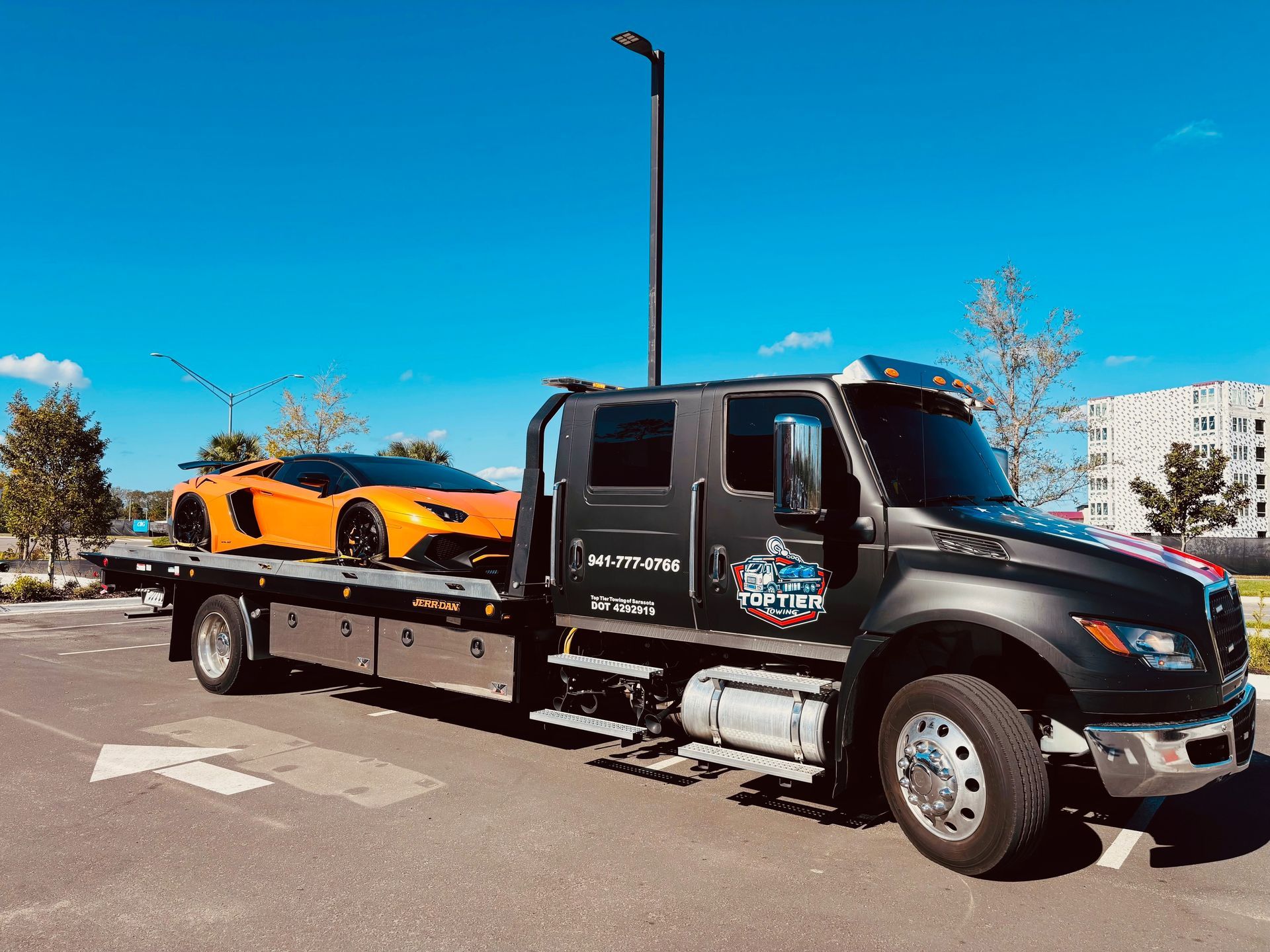 A black tow truck transporting a bright orange Lamborghini sports car in a paved parking lot under a blue sky.