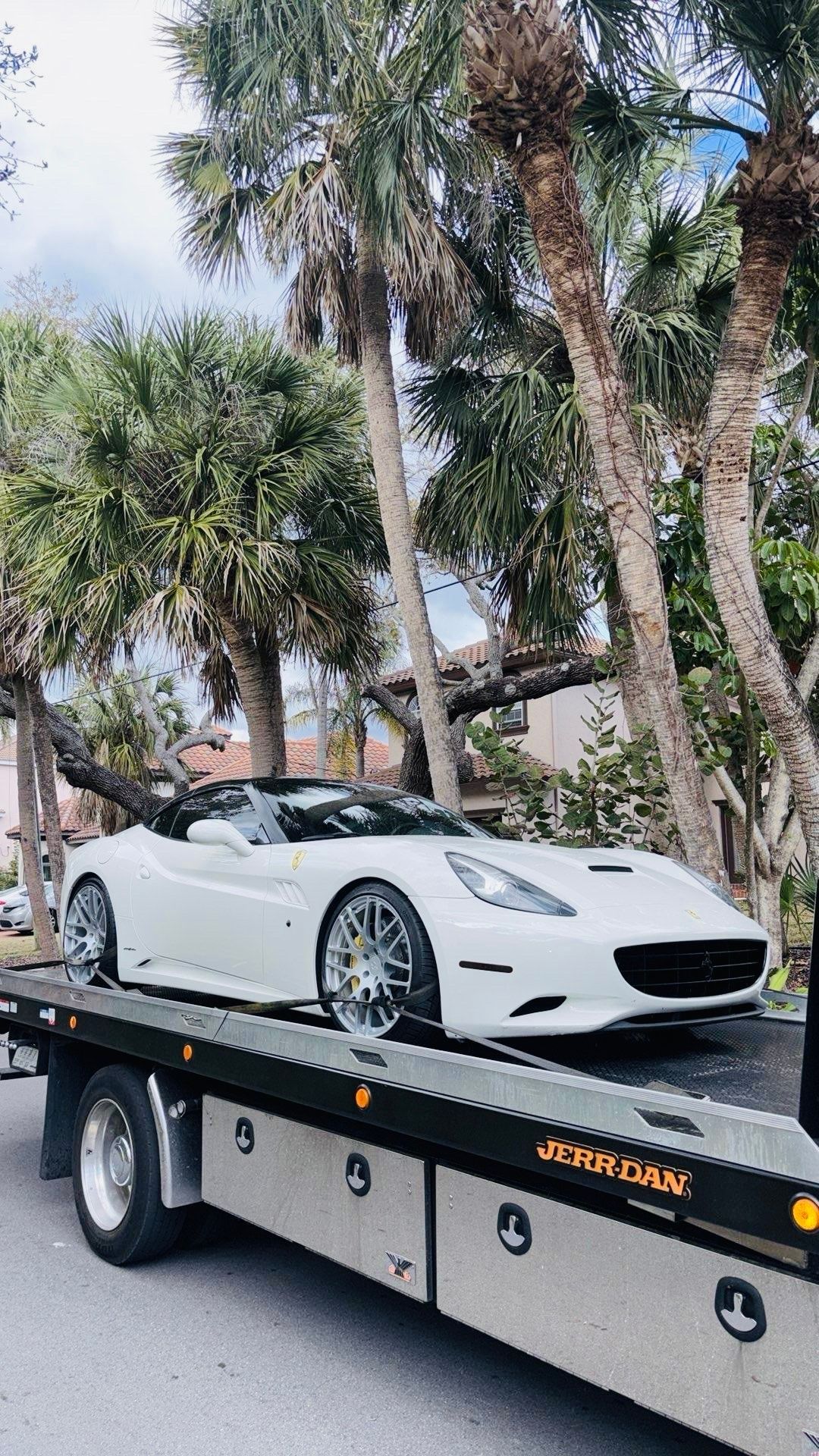 A white convertible Ferrari parked on the bed of a tow truck, framed by palm trees on a sunny day.