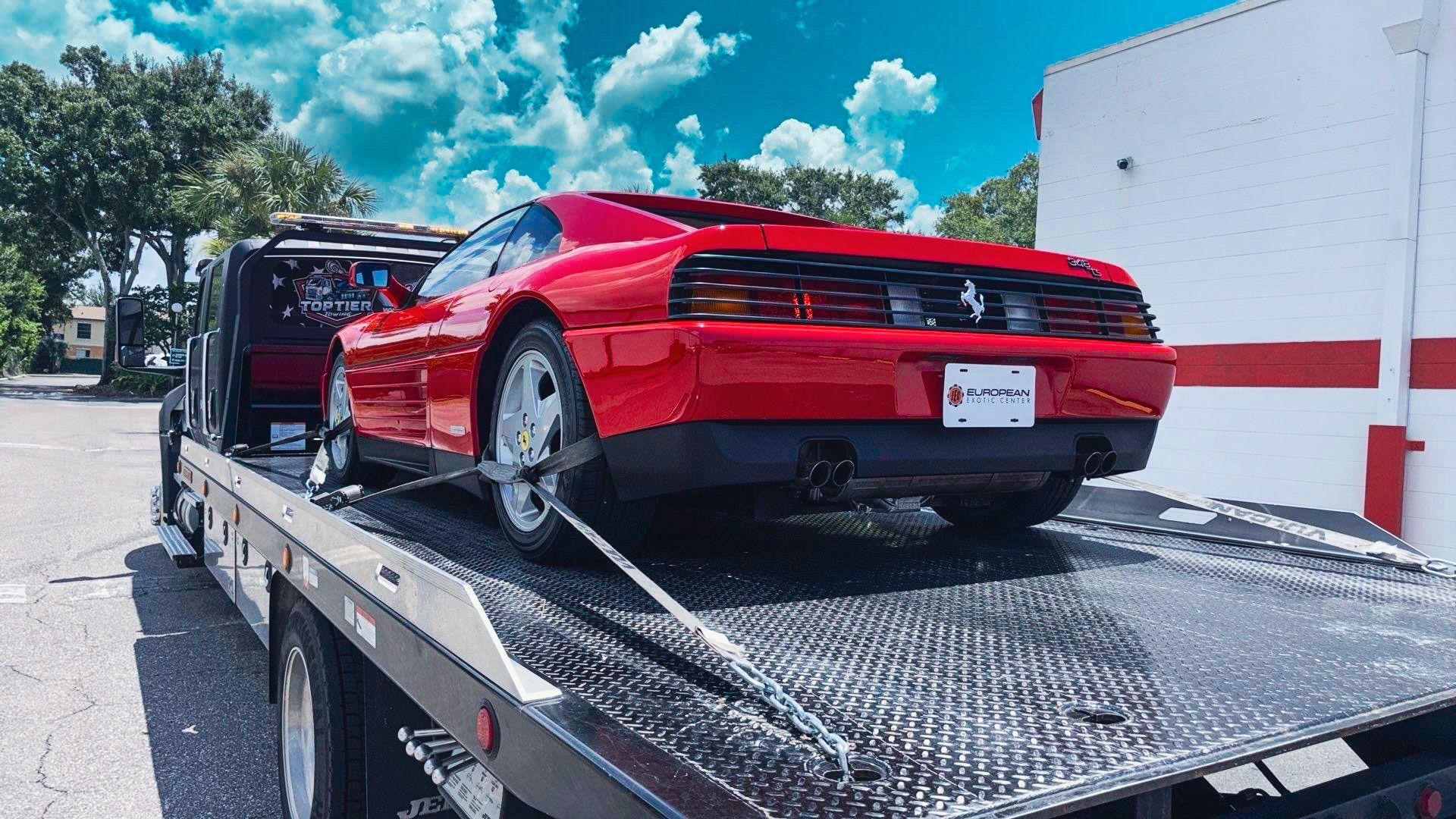 A red Ferrari sports car secured by straps on the back of a flatbed tow truck in a sunny outdoor parking lot.