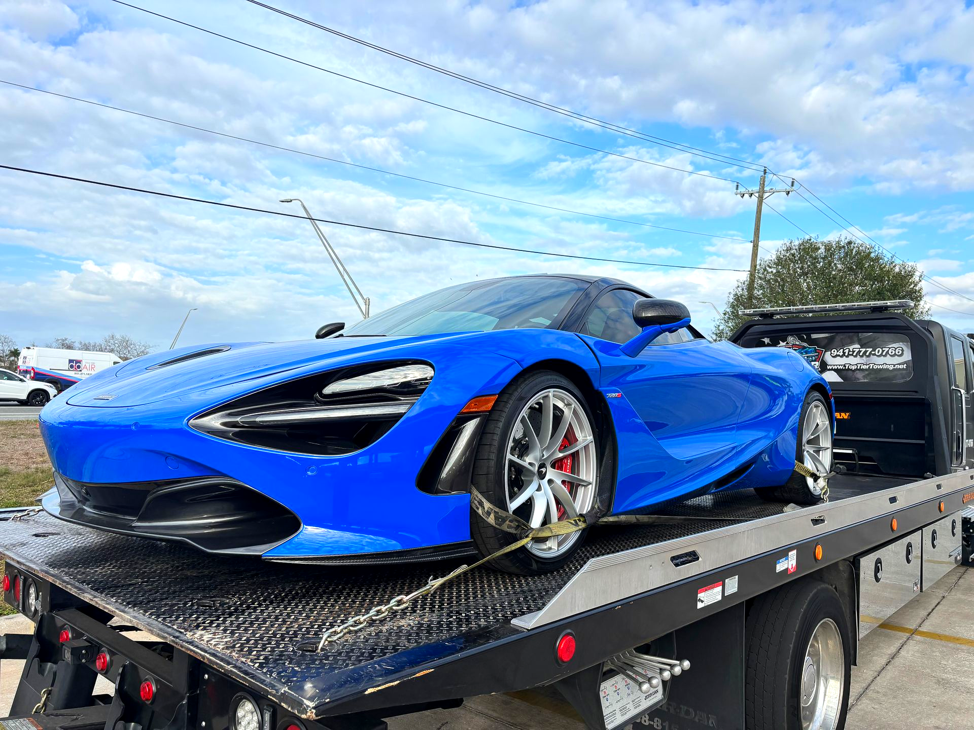 A bright blue McLaren supercar secured with straps on the flatbed of a tow truck against a cloudy sky.