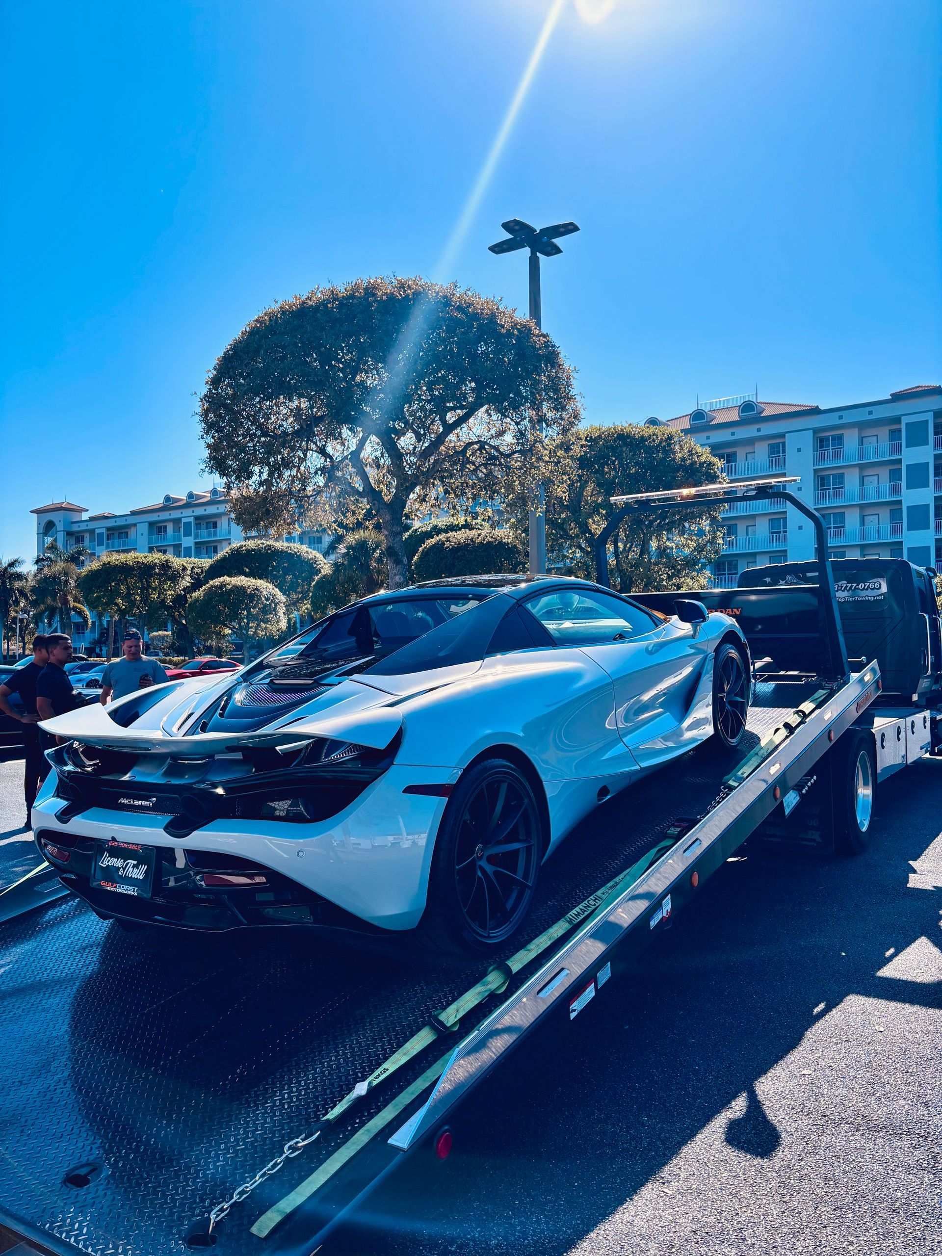A white McLaren sports car being winched onto the back of a flatbed tow truck on a sunny day.