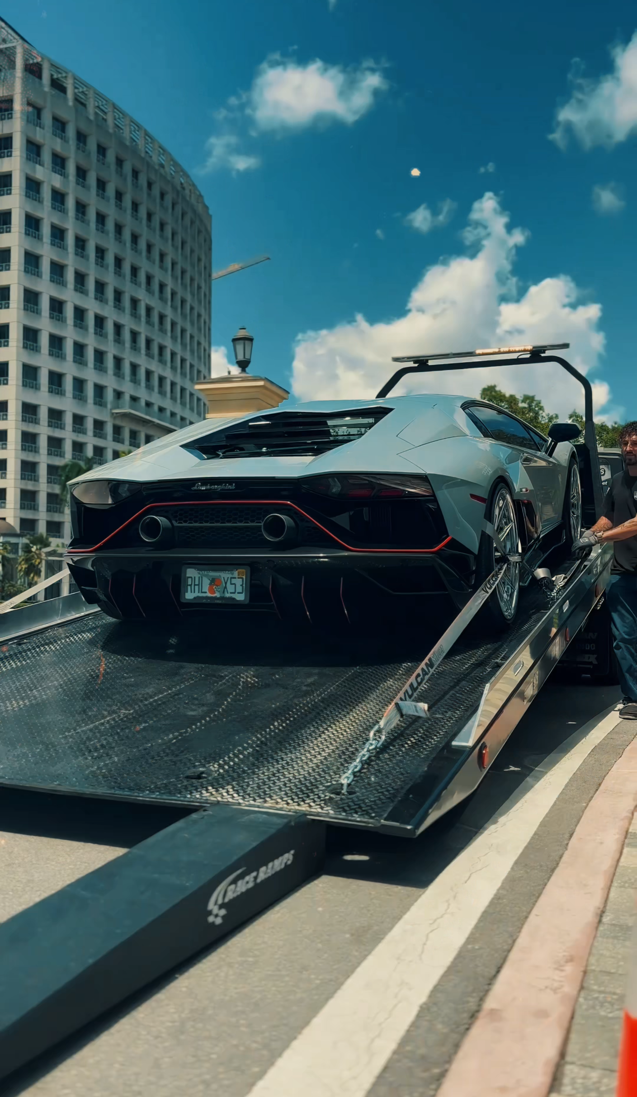 A light blue Lamborghini Aventador being winched onto a flatbed tow truck on a sunny city street.