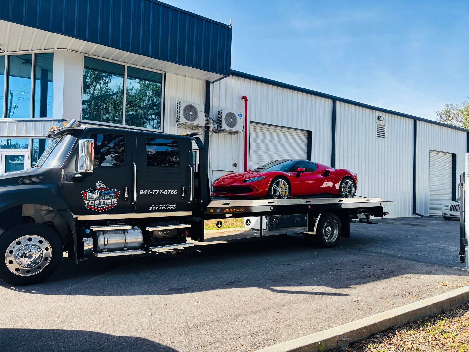 A black tow truck with a red sports car loaded on its flatbed parked outside a warehouse building on a sunny day.