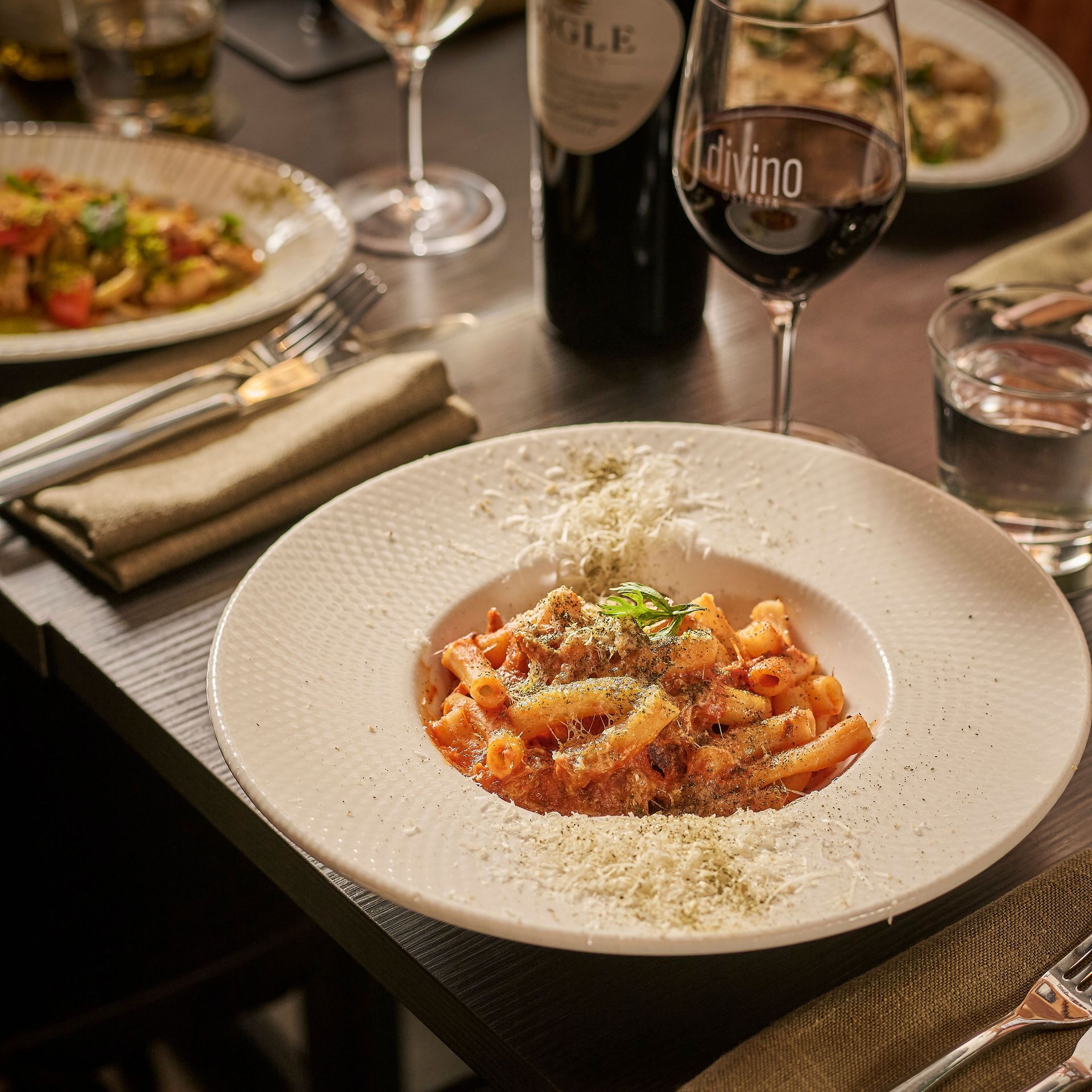 A plated pasta dish with sauce and grated cheese on a restaurant table with wine glasses.