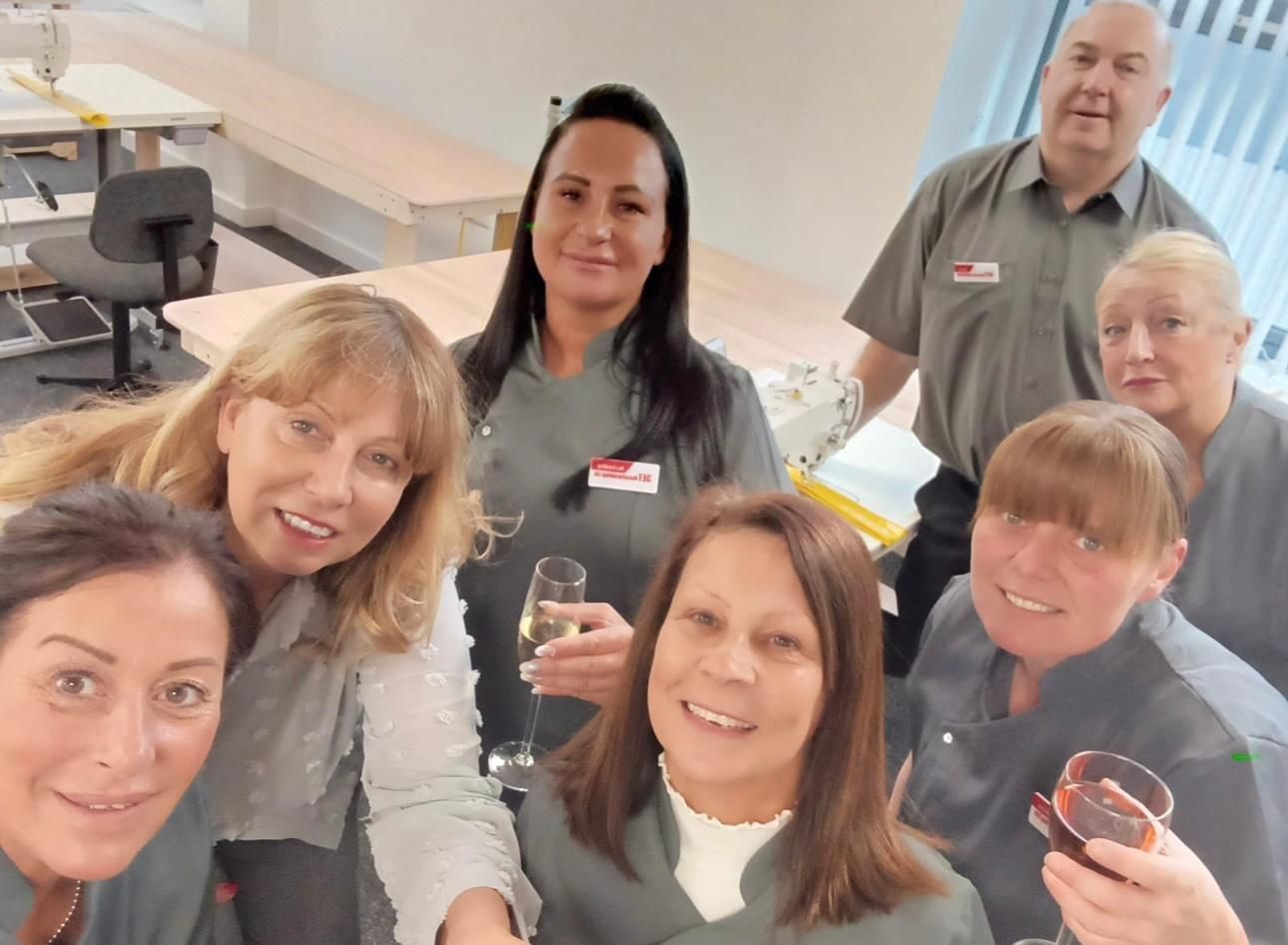 Group of people in gray uniforms with drinks, smiling in a room with long tables.