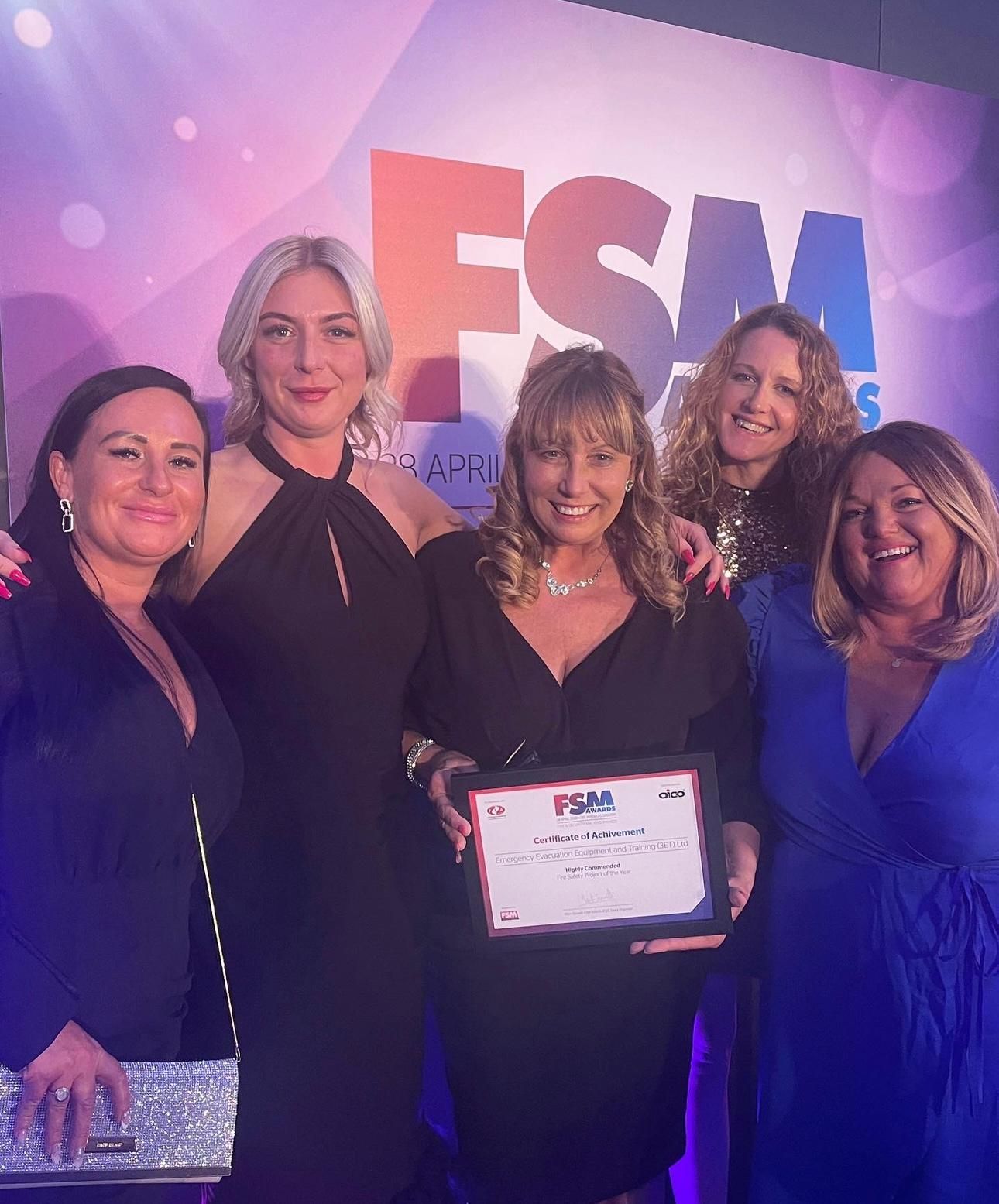Five women smiling, holding an award, against a colorful backdrop with 