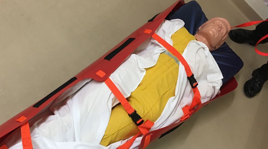 Mannequin on a red stretcher, secured with orange straps. Indoors, likely a training exercise.