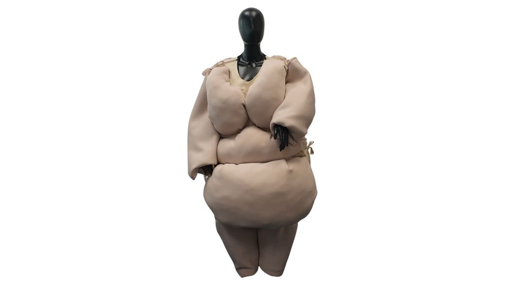 Mannequin in beige, padded suit, mimicking a large body, with dark head and hands, against white.