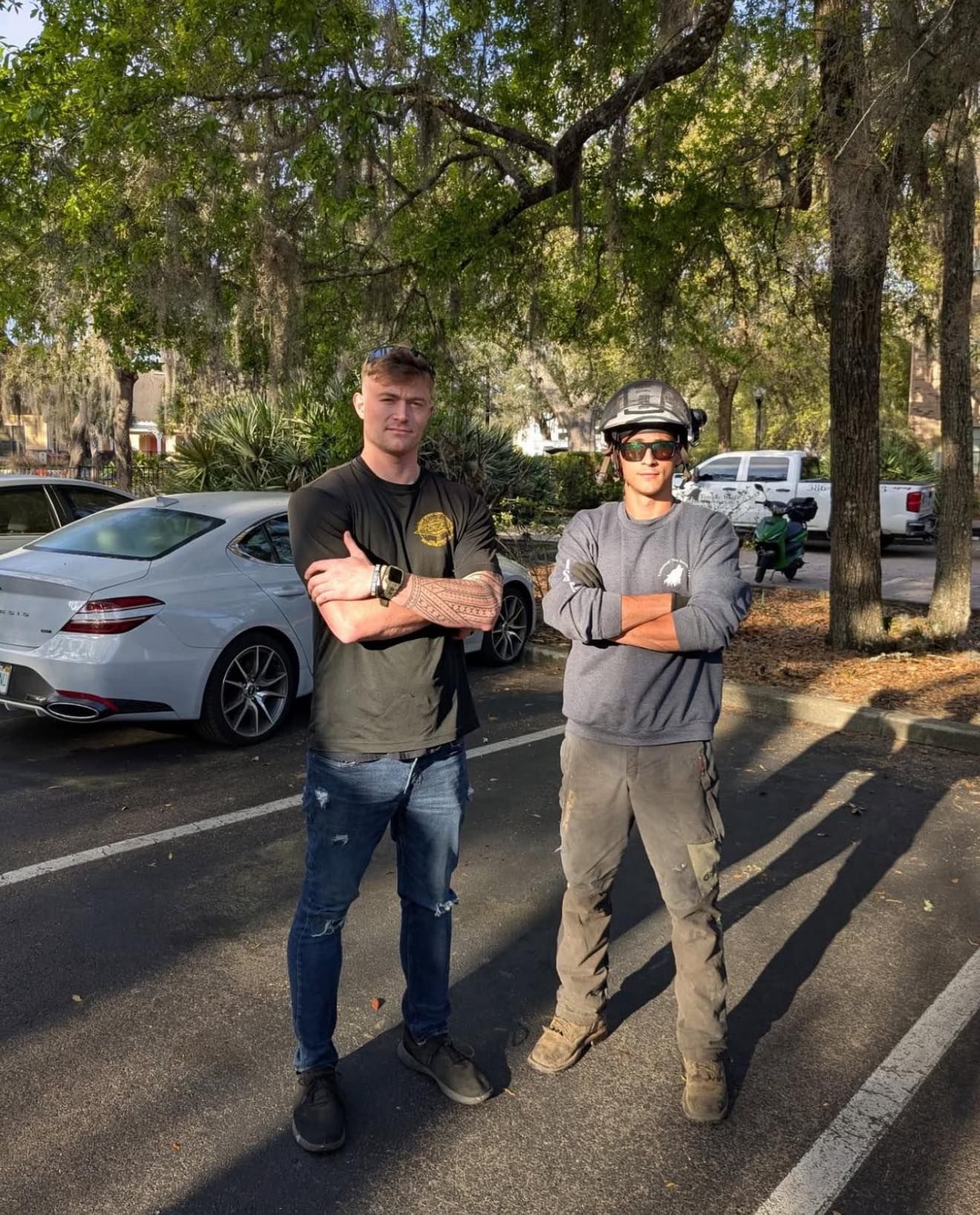 Two men with arms crossed stand outdoors. One wears a dark t-shirt and jeans; the other, a long-sleeve, and work pants.