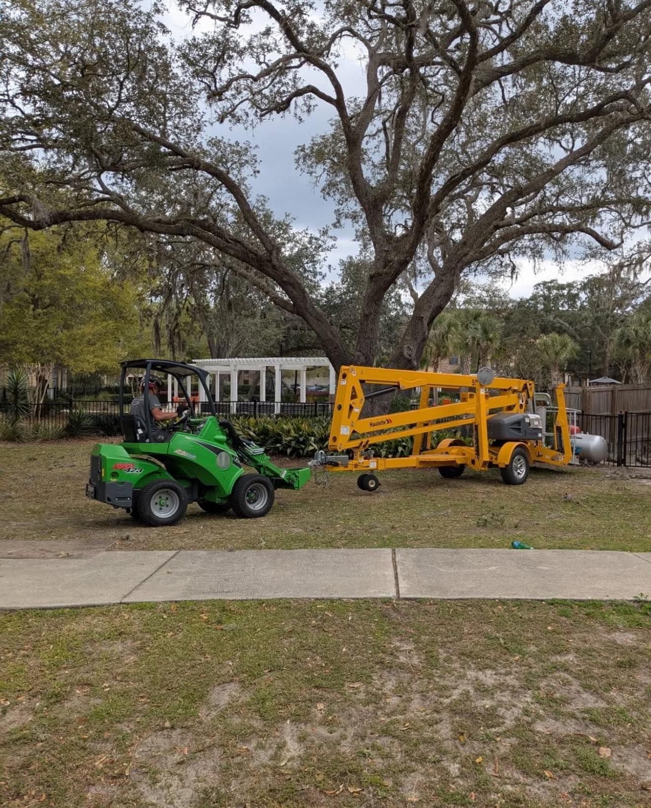 Green tractor towing a yellow lift on a grassy lawn under a large tree; a person is driving.