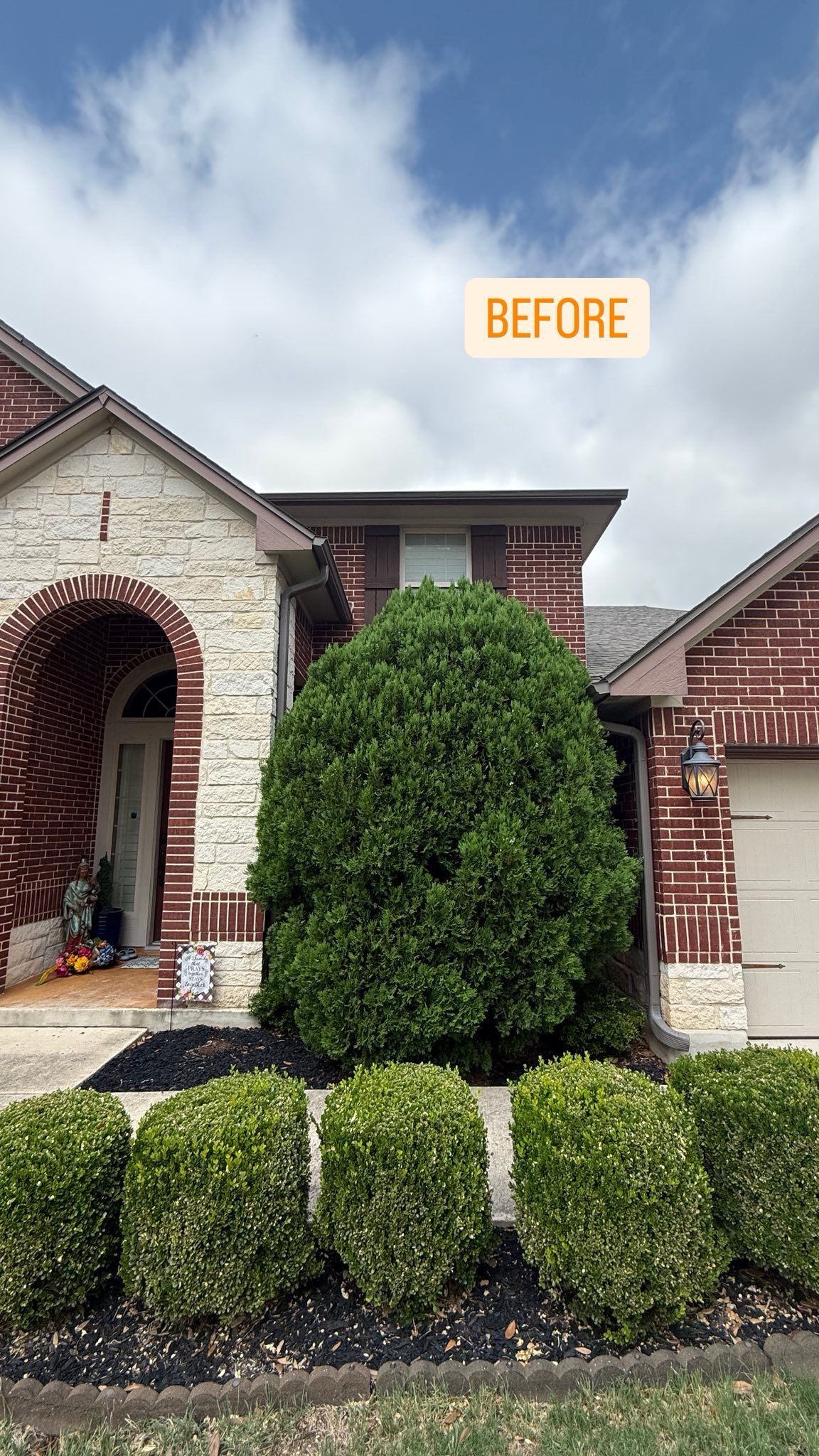 House exterior, before landscaping. Brick and stone facade with large green bush and trimmed shrubbery.