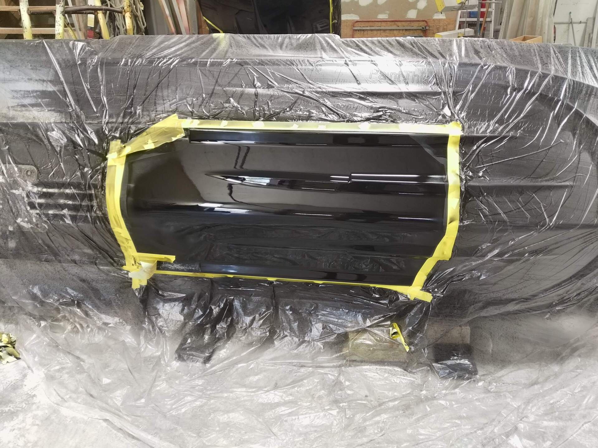 Black car panel masked with yellow tape and plastic sheeting in a workshop