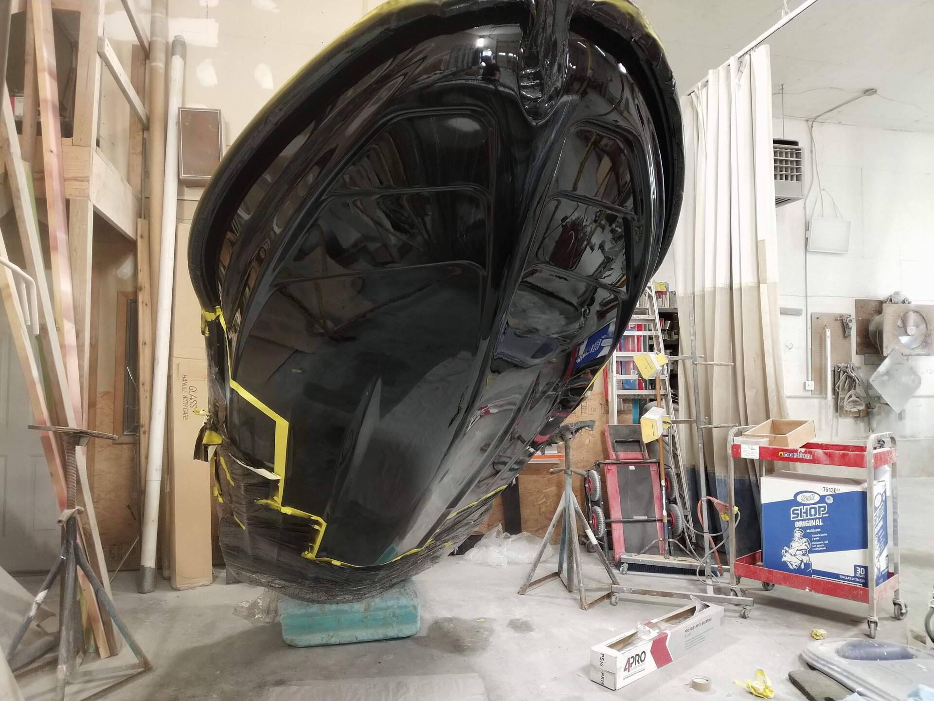 Large glossy black boat hull in a workshop, propped on supports with tools and ladders nearby.