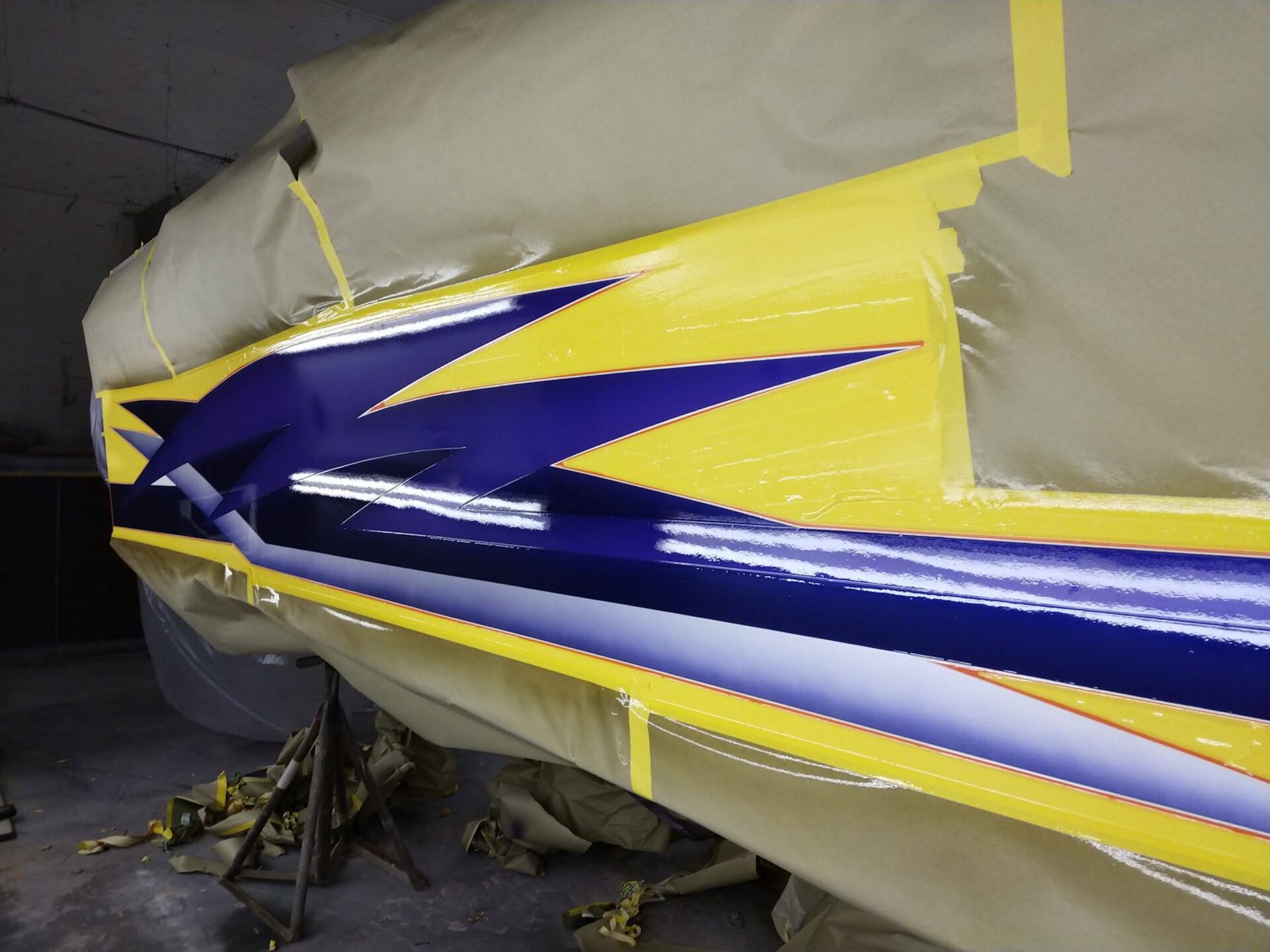 Boat hull being painted with blue, white, and yellow racing stripes in a workshop
