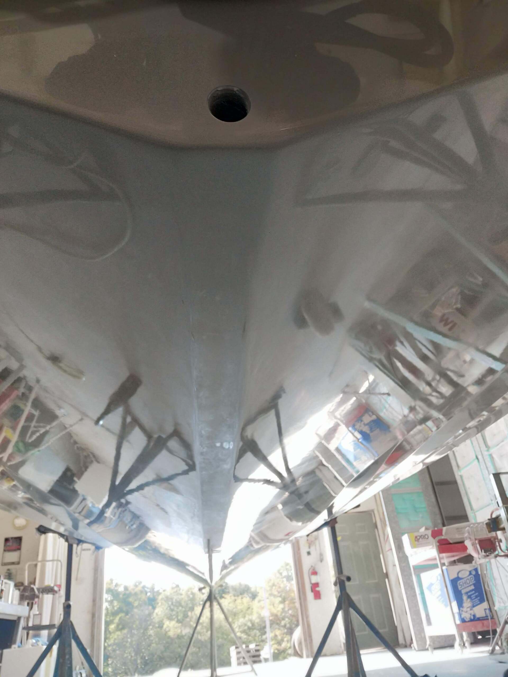 Inside a hangar, a white boat hull is shown from below, with support stands and equipment on both sides.