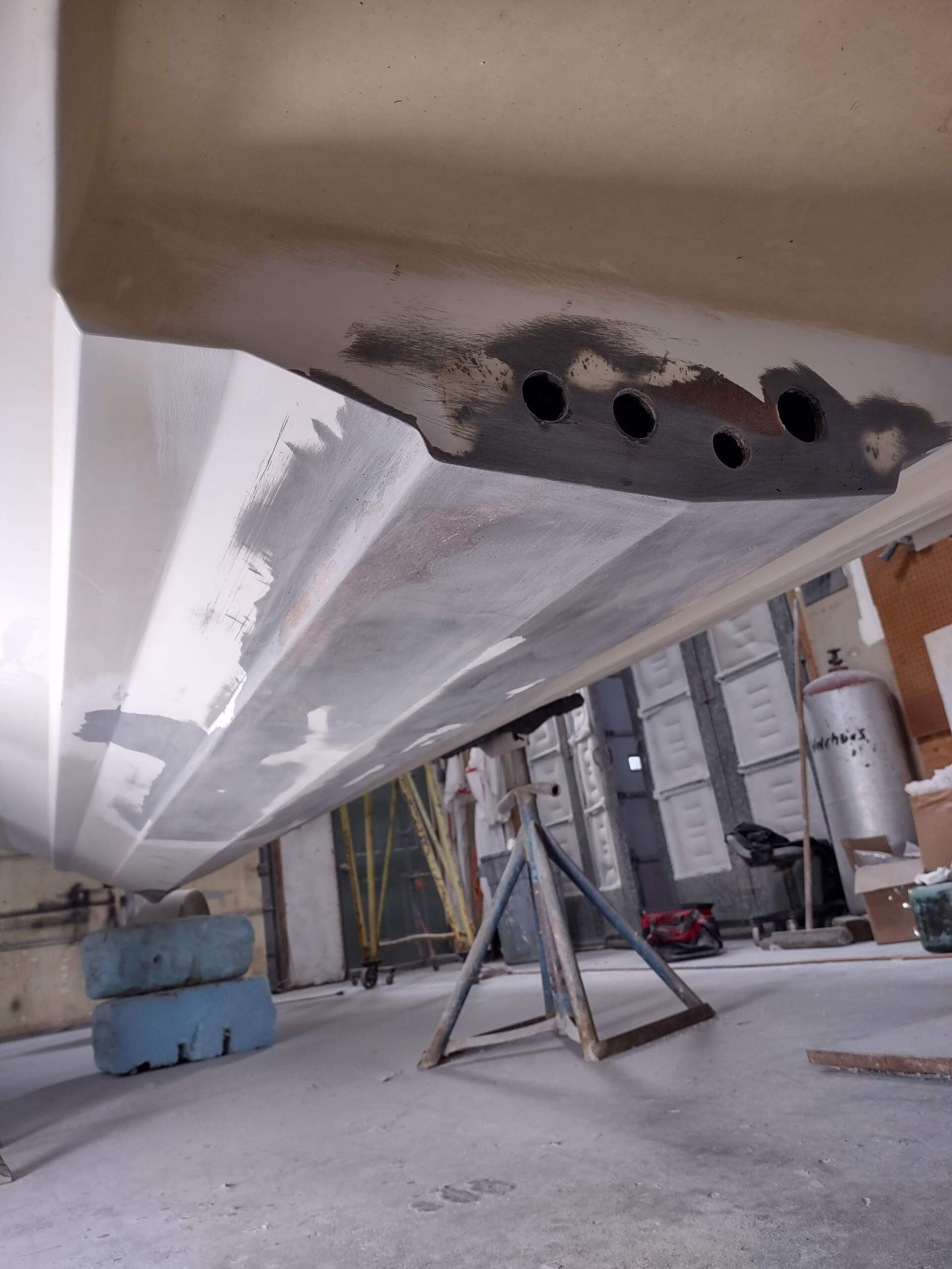 Underneath a boat hull in a workshop, showing a gray underside with holes and support stands on a concrete floor