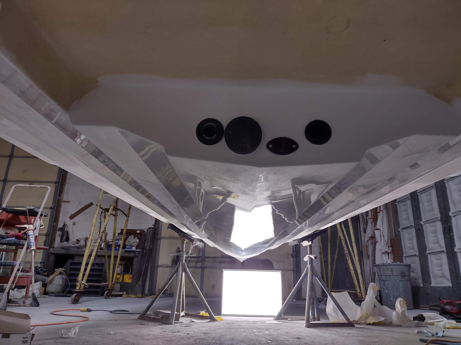 Underneath a boat hull in a workshop, showing circular openings and support stands.