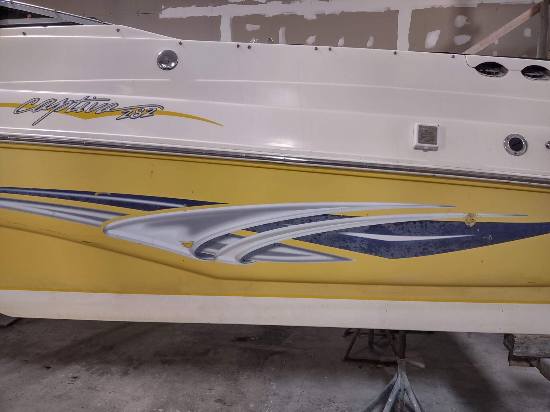 Side view of a white and yellow boat hull with gray and blue decorative stripes inside a garage.